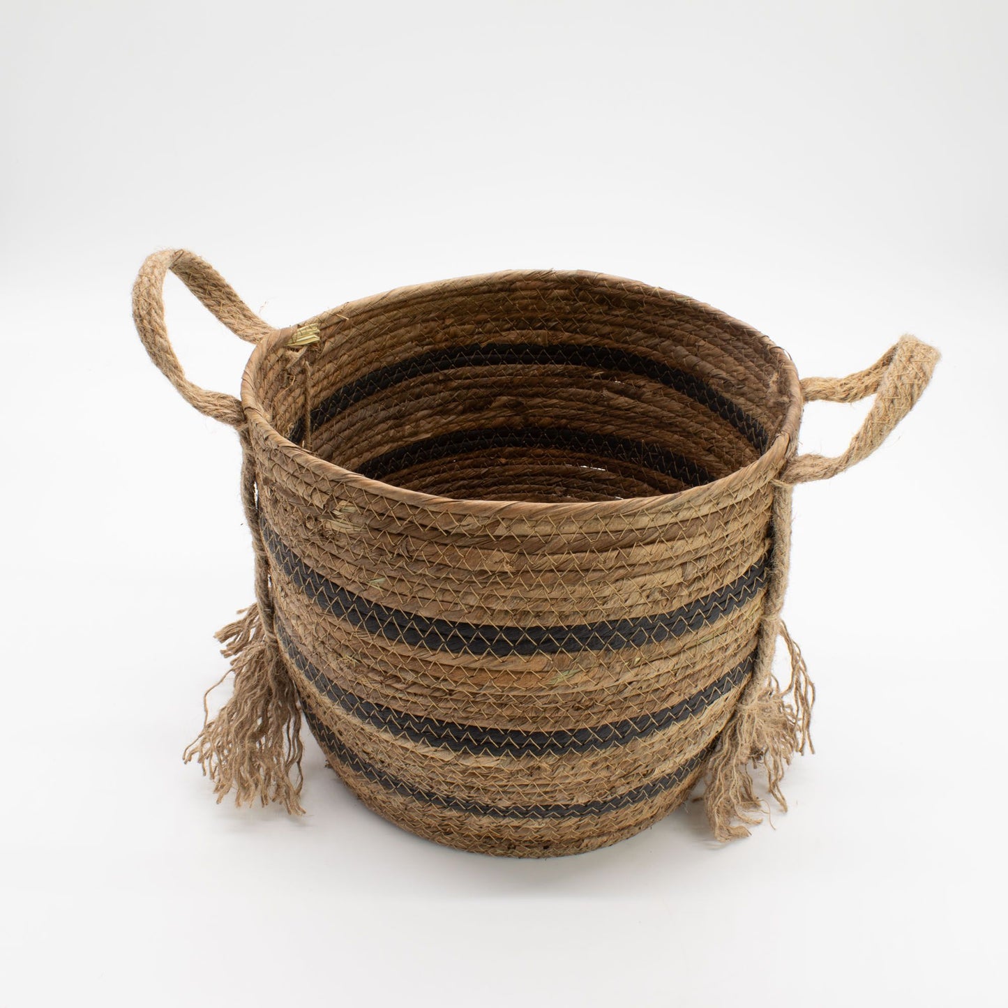 Cesta redonda de hoja de plátano y pasto marino trenzado, color natural y bandas negras con asas y flecos decorativos.