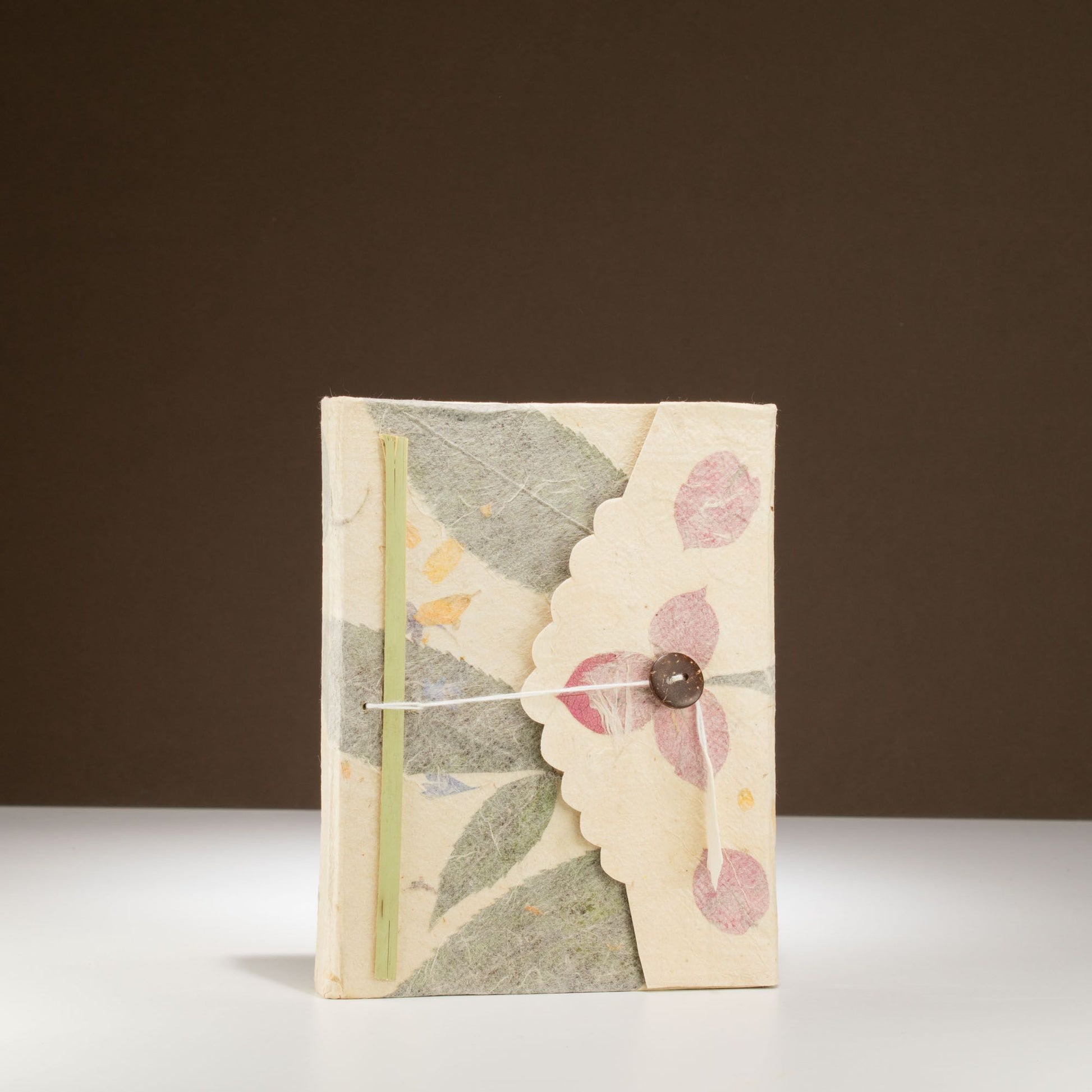 Cuaderno artesanal de papel reciclado con flores rosadas, tapa en tonos crema y cinta de cierre con botón.