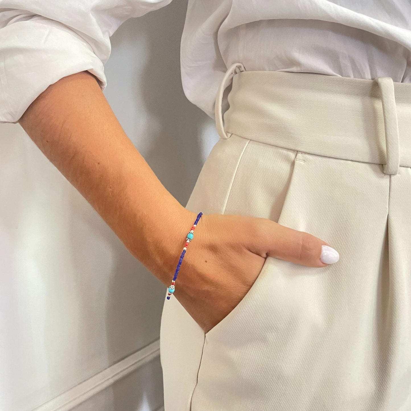 Pulsera de cuentas azul y blanca con detalles en rojo, estilo bohemio, sobre blusa blanca y pantalones color crema.