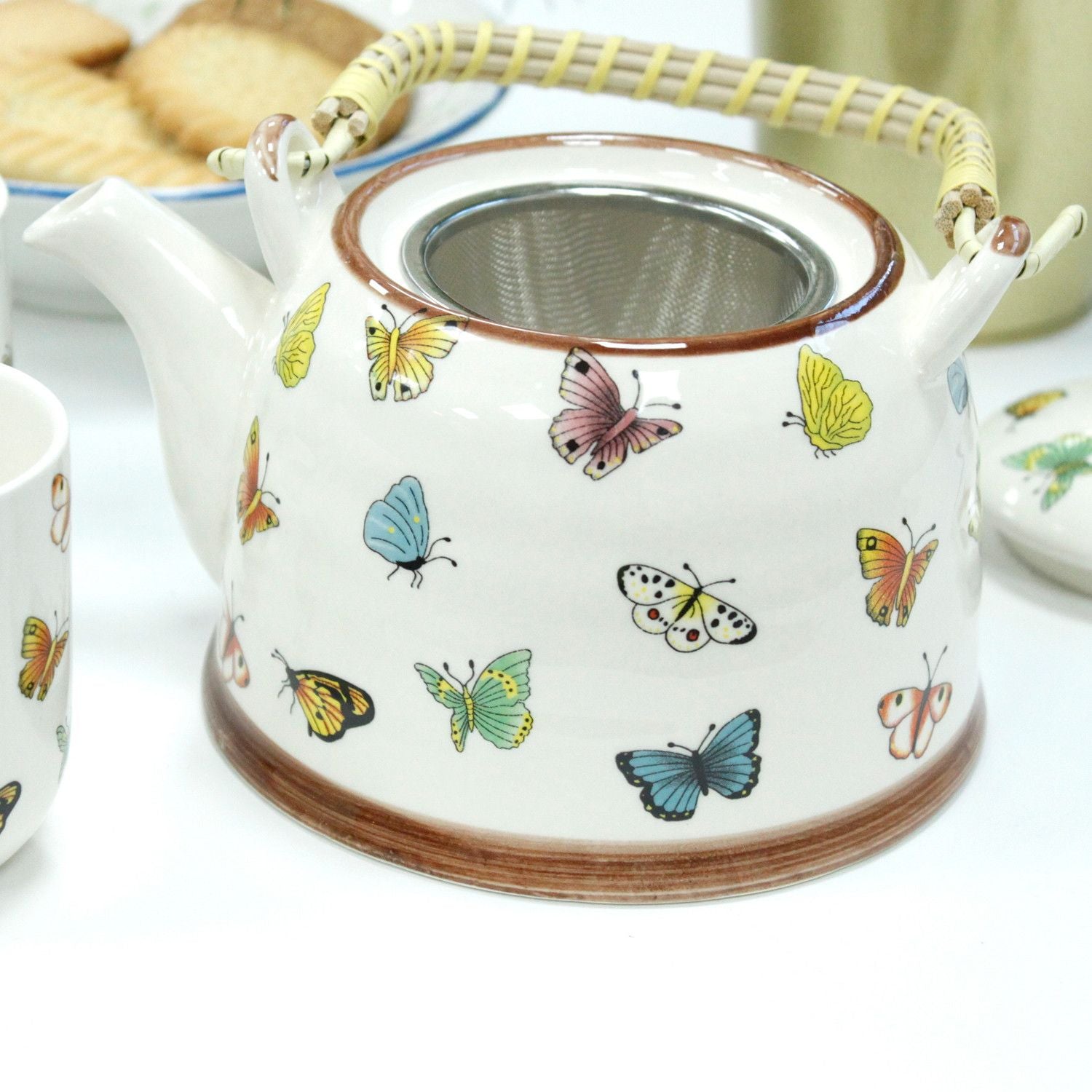 Tetera de cerámica blanca con mariposas multicolores y asa de mimbre, estilo vintage.