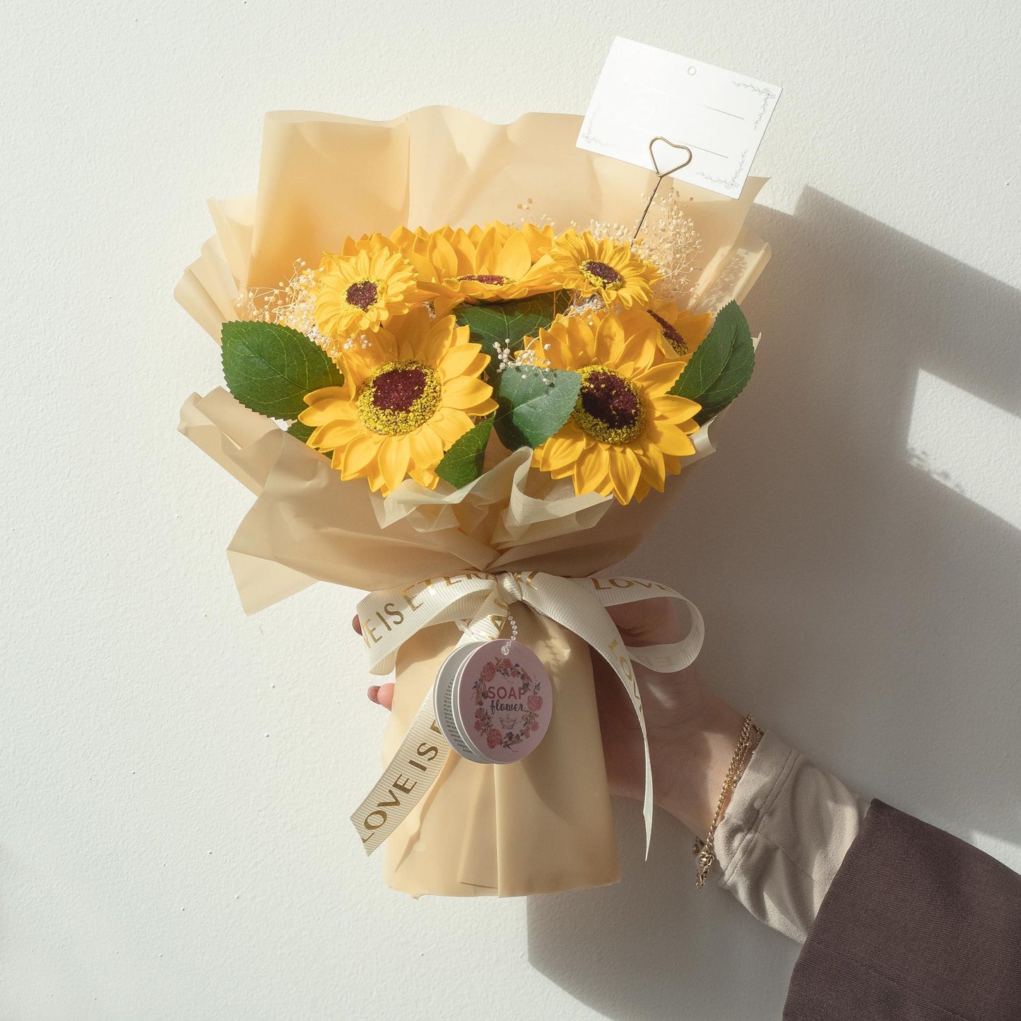 Ramo de flores de jabón con girasoles amarillos, hojas verdes y envoltura beige, decorado con una cinta blanca.