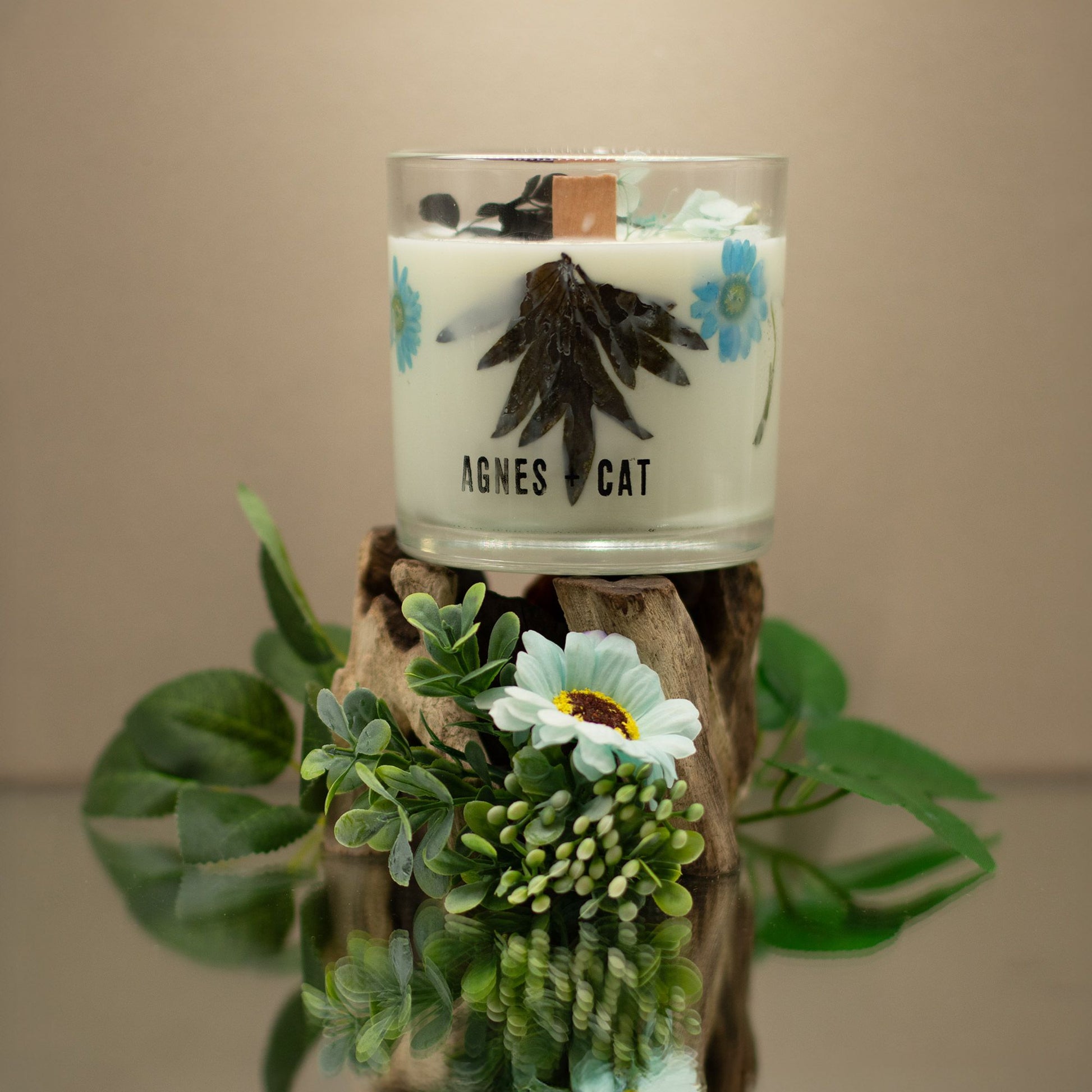 Vela aromática blanca con flores azules y madera, presentada en envase de vidrio, estilo natural y elegante.