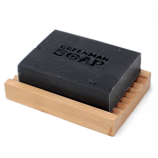Pastilla de jabón negra rectangular sobre una jabonera de madera clara con diseño estriado sobre fondo blanco