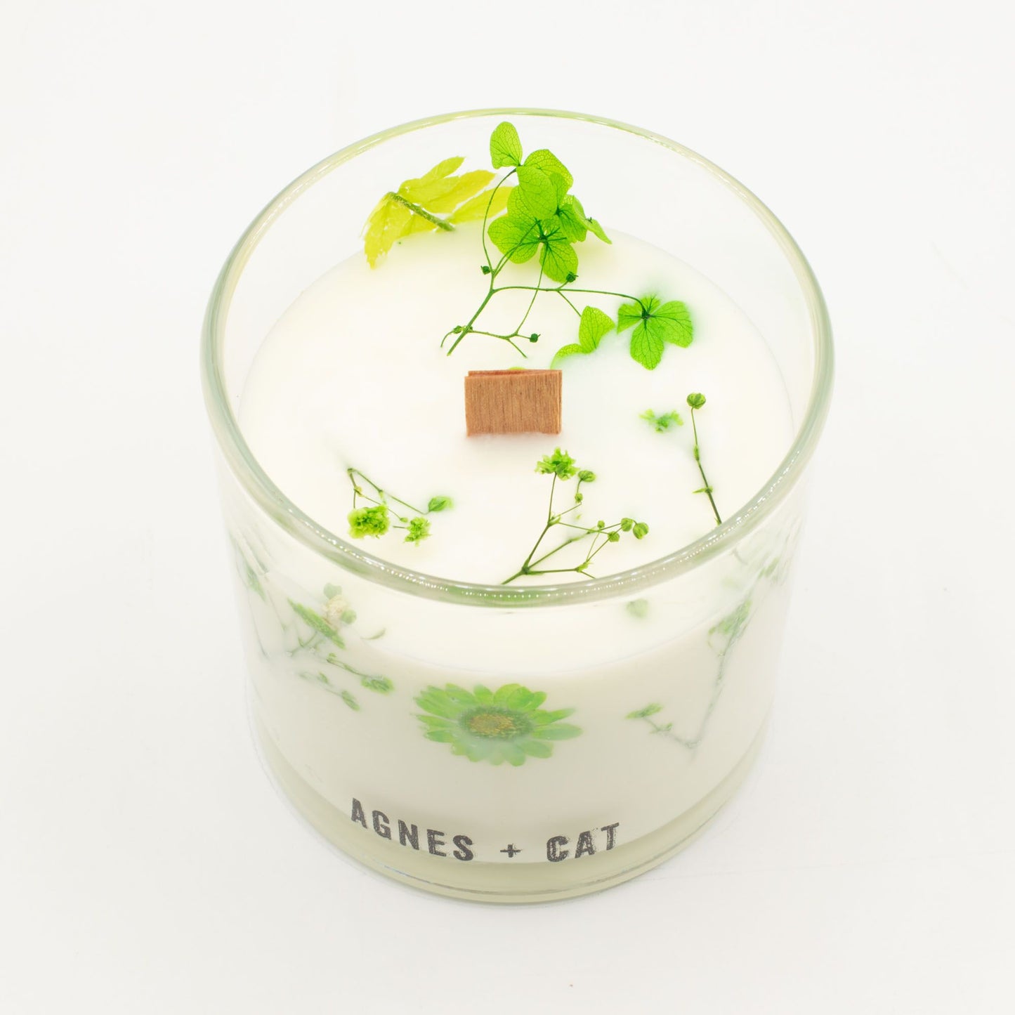 Vela decorativa blanca en frasco de vidrio con flores verdes y tapa de madera, estilo elegante y natural.