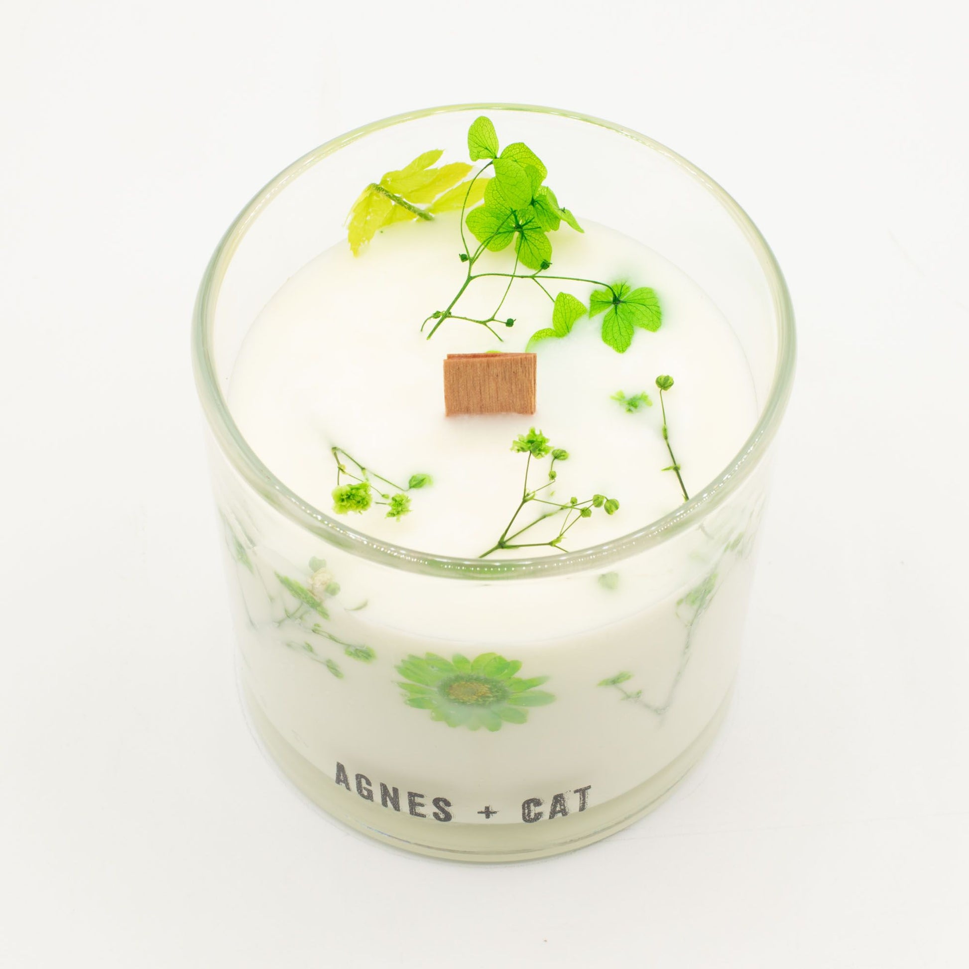 Vela decorativa blanca en frasco de vidrio con flores verdes y tapa de madera, estilo elegante y natural.