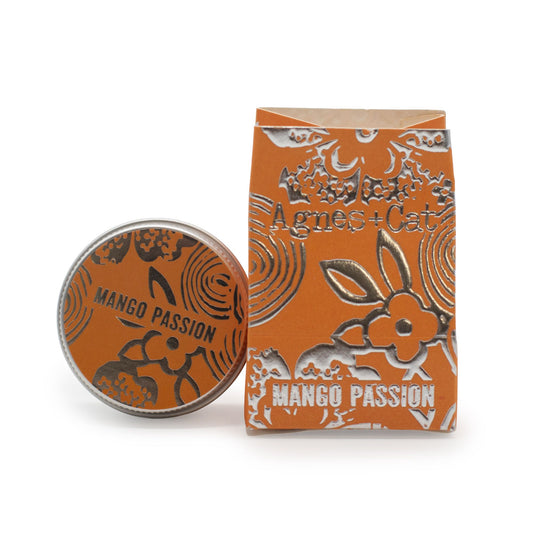 Bálsamo labial Agnes + Cat Mango Passion: caja de cartón y lata redonda, fondo naranja con detalles florales plateados.