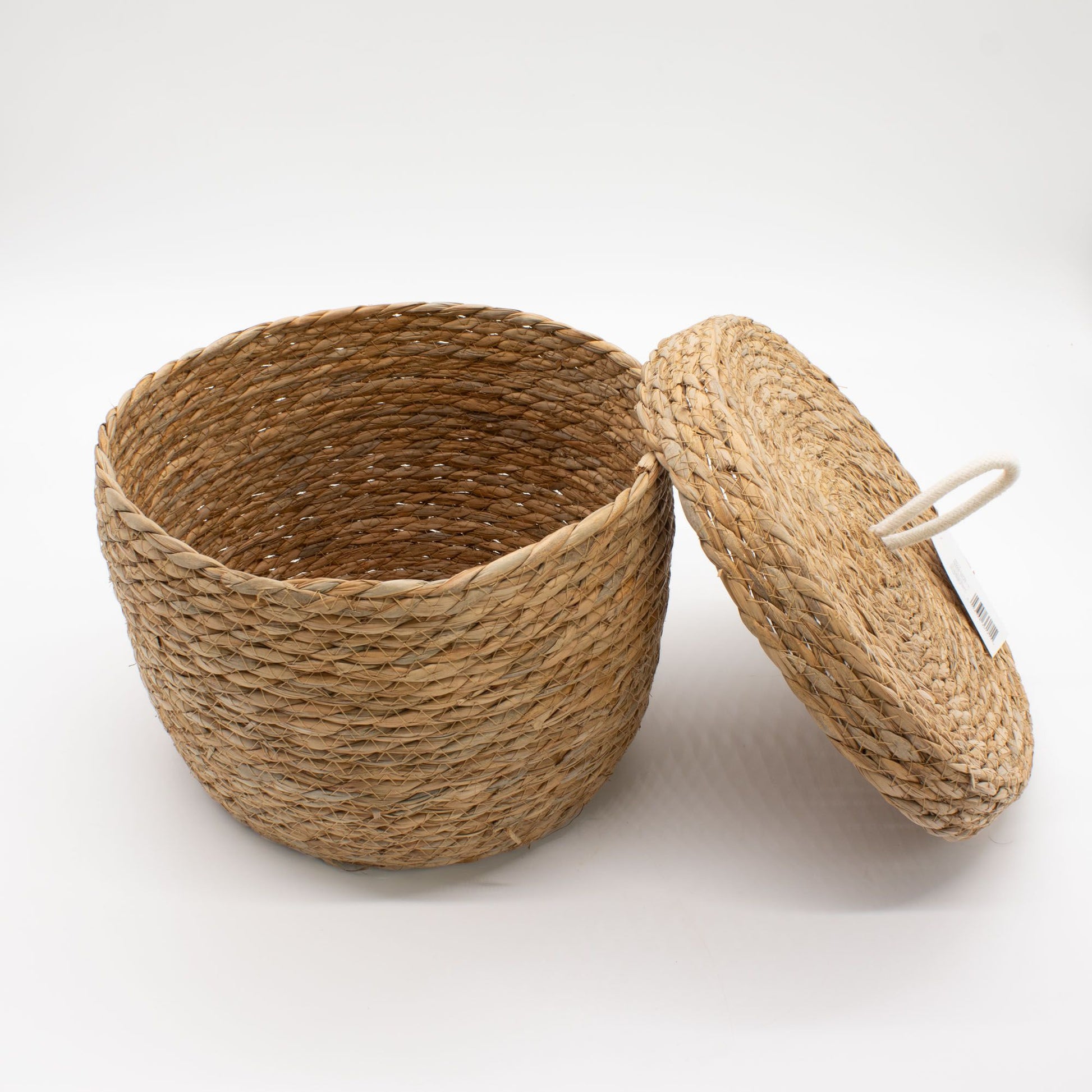 Cesta redonda tejida de hierba marina color natural con tapa y asa de cuerda blanca sobre fondo blanco.
