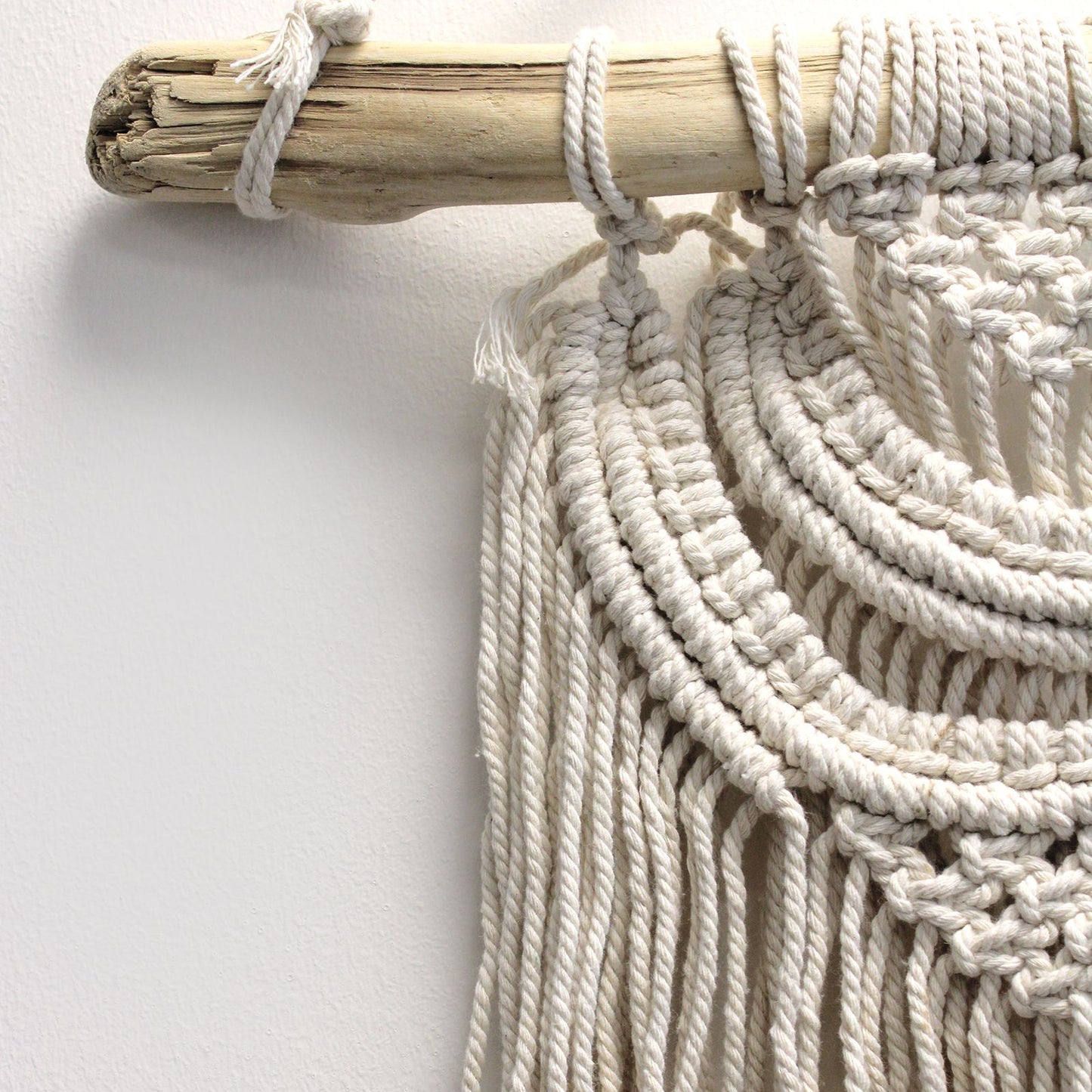 Pared decorativa de macramé beige, hecha de hilo de algodón, estilo bohemio con soporte de tronco natural.