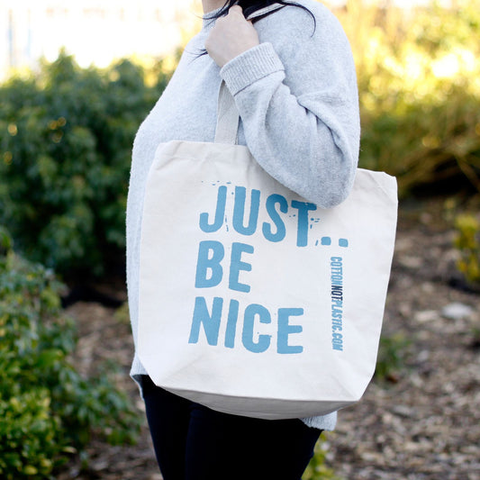 Bolsa de algodón color beige con el texto en azul "JUST BE NICE" colgada al hombro sobre un fondo de jardín.