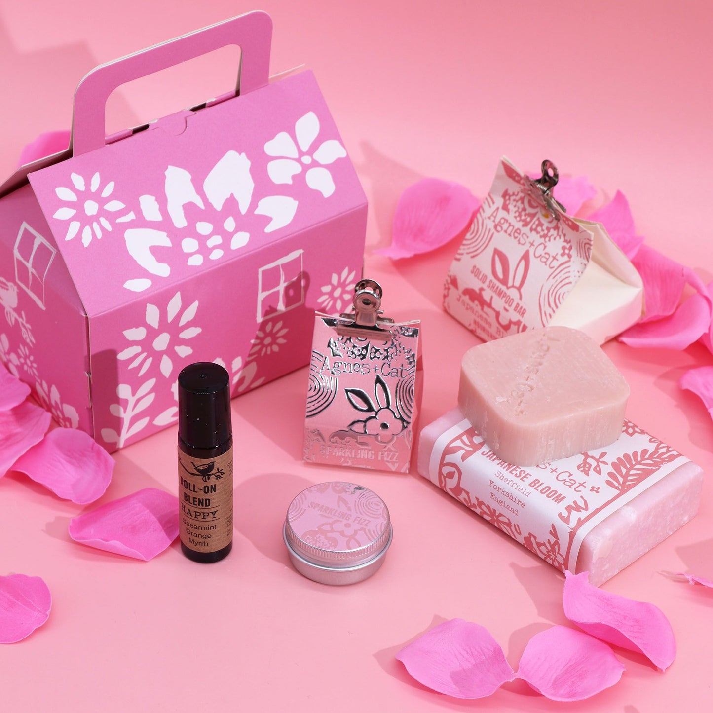 Set de regalo Agnes & Cat en caja rosa con pétalos, jabones, champú sólido, aceite roll-on y lata sobre fondo rosa.