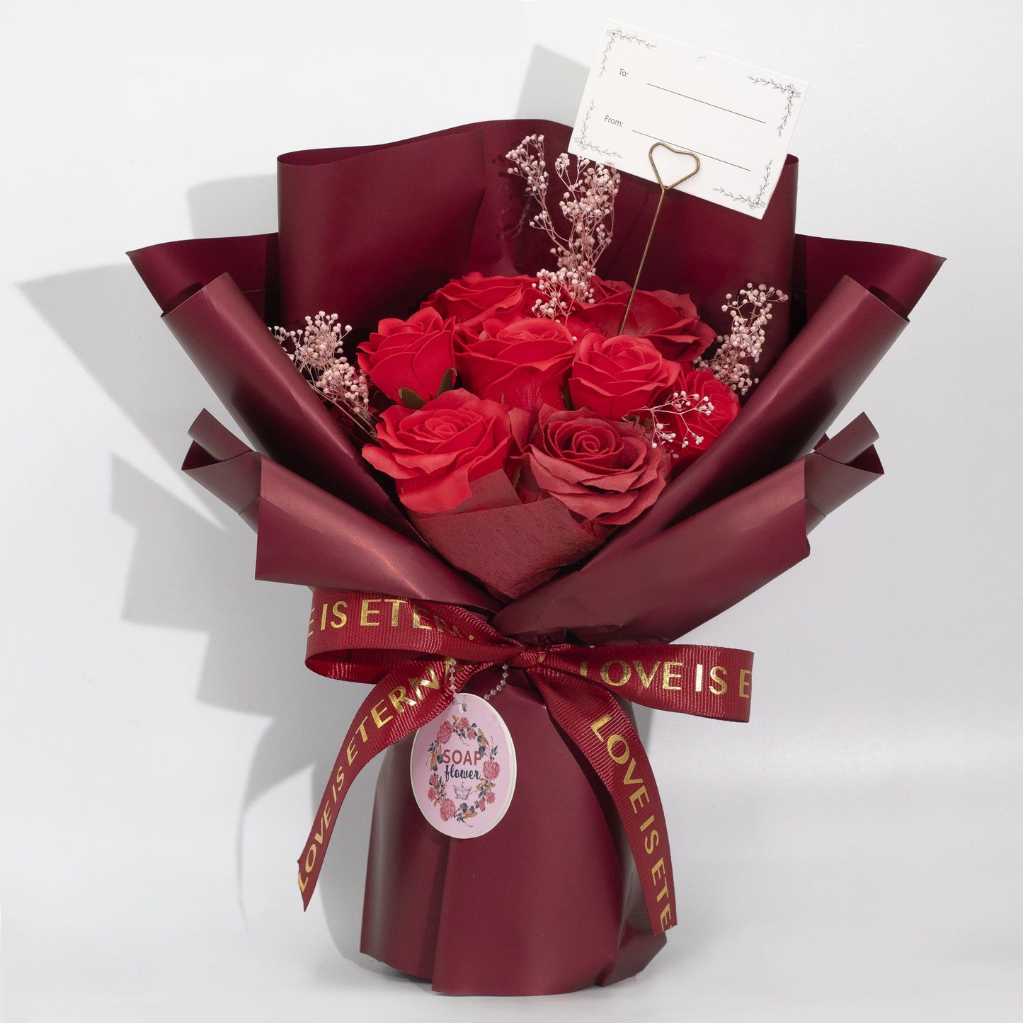 Ramo de rosas rojas en envoltura burdeos, con tarjeta y cinta dorada "Love Is Eternal". Ideal para regalos románticos.