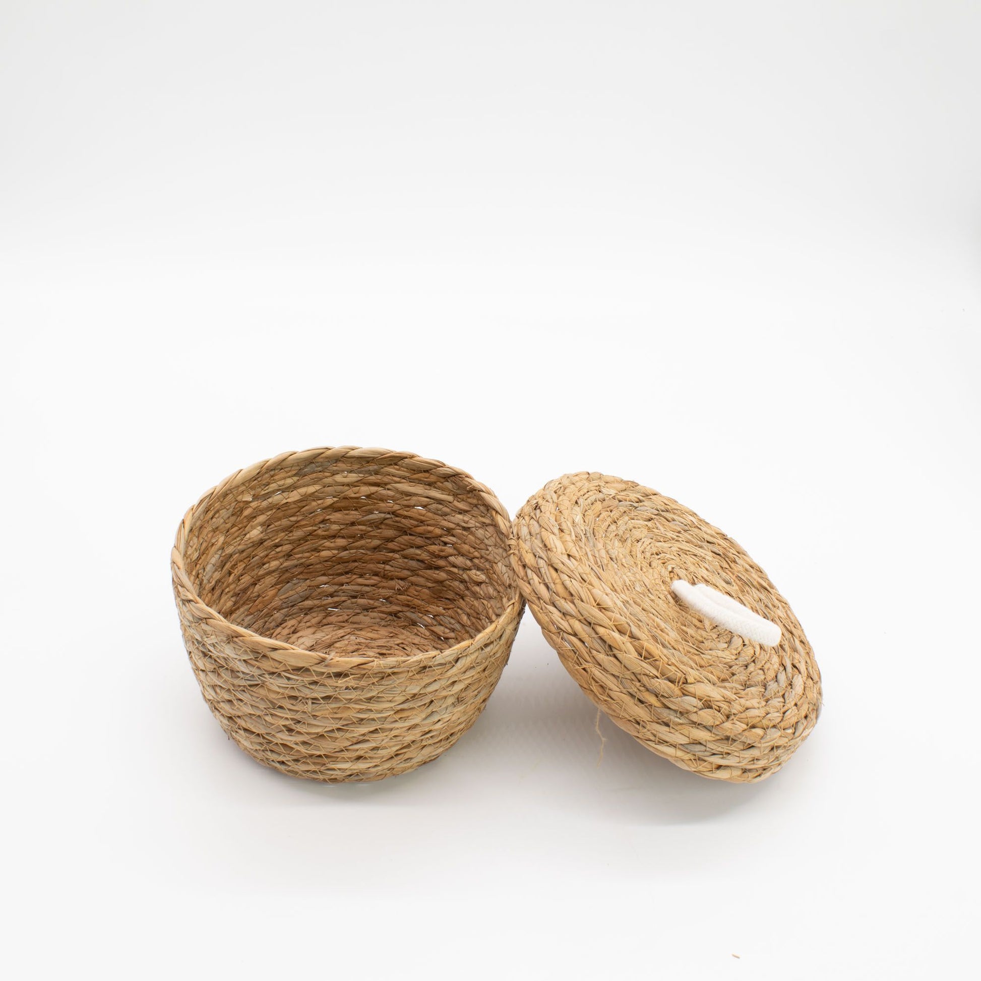 Cesta redonda de hierba marina trenzada con tapa y asa de algodón blanco sobre fondo blanco. Estilo natural y artesanal.