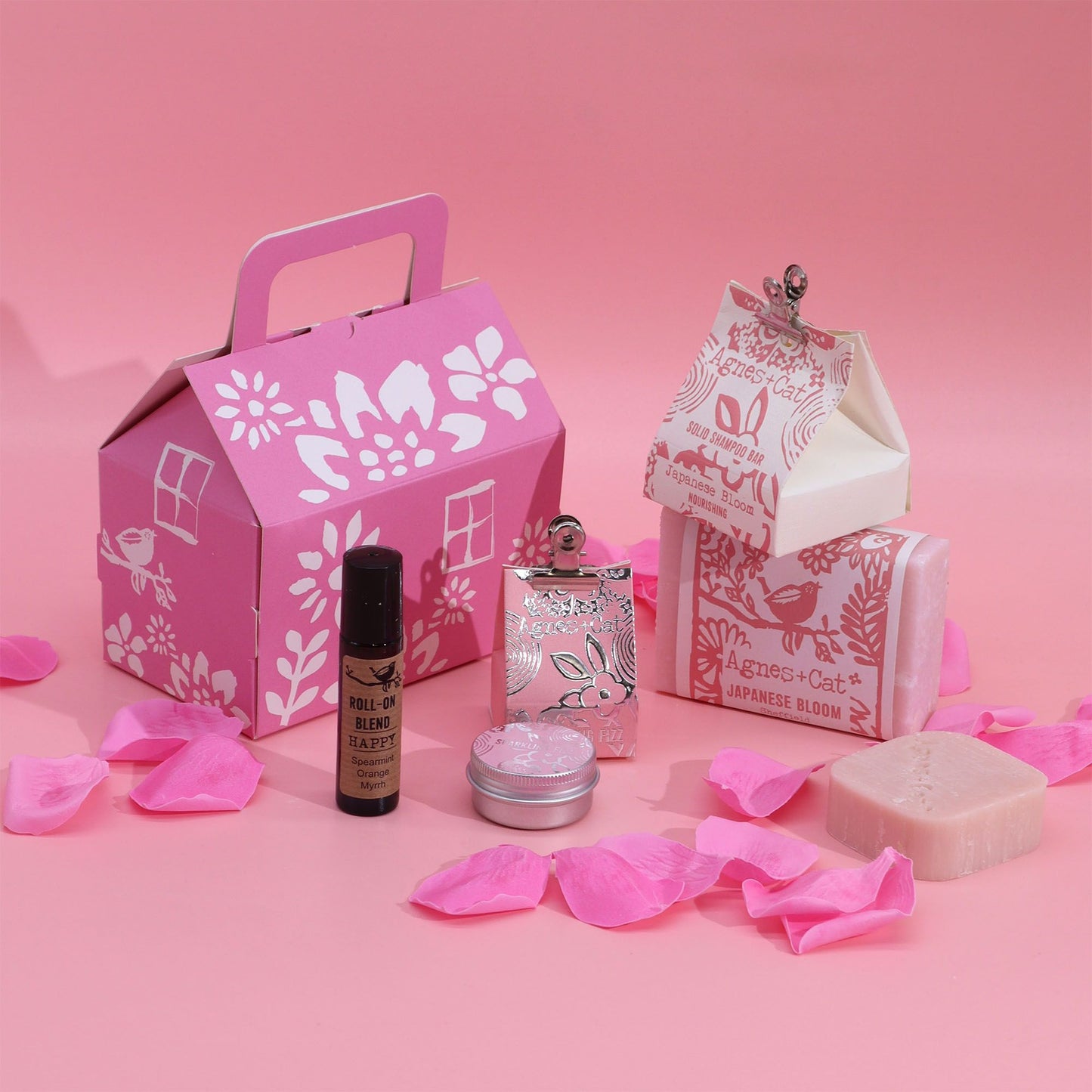 Set de regalo Agnes & Cat: caja tipo casa rosa, jabón, champú, roll-on y lata sobre fondo rosa con pétalos decorativos.
