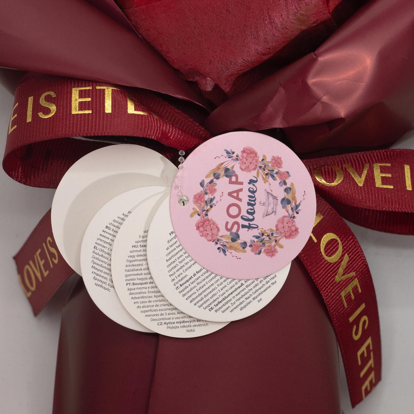 Ramo de flores de jabón en tonos rosa y rojo, con cinta roja brillante y tarjeta de información. Elegante y decorativo.