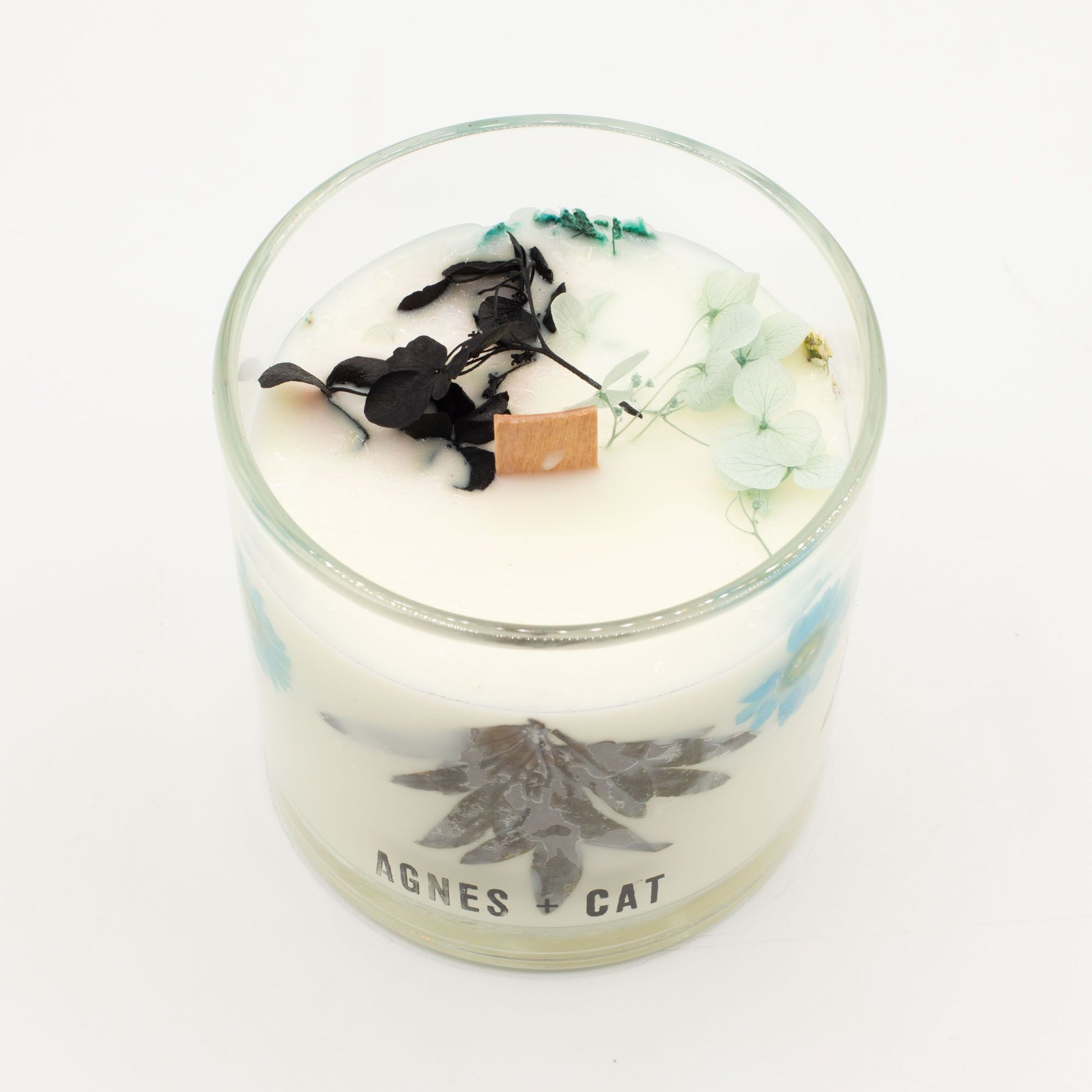 Vela aromática blanca con flores secas, detalles en azul y tapa de madera, estilo elegante y natural.