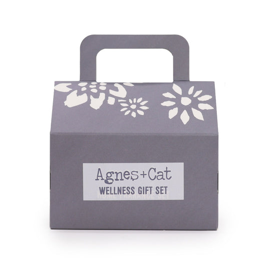Caja de regalo gris con asa, detalles florales blancos y etiqueta central "Agnes + Cat Wellness Gift Set".