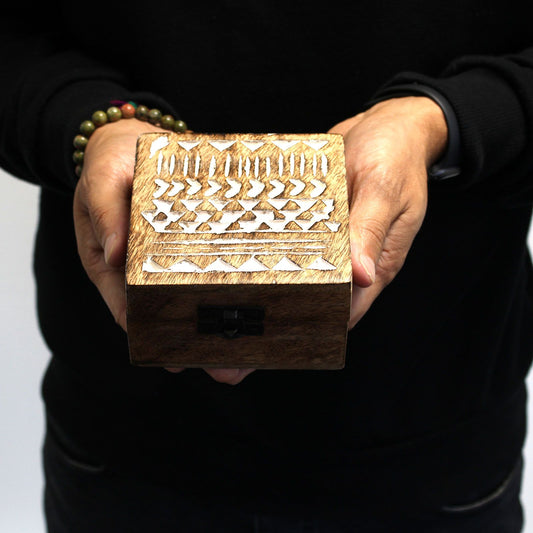 Caja cuadrada de madera oscura con grabados geométricos blancos tipo azteca, sostenida por unas manos.