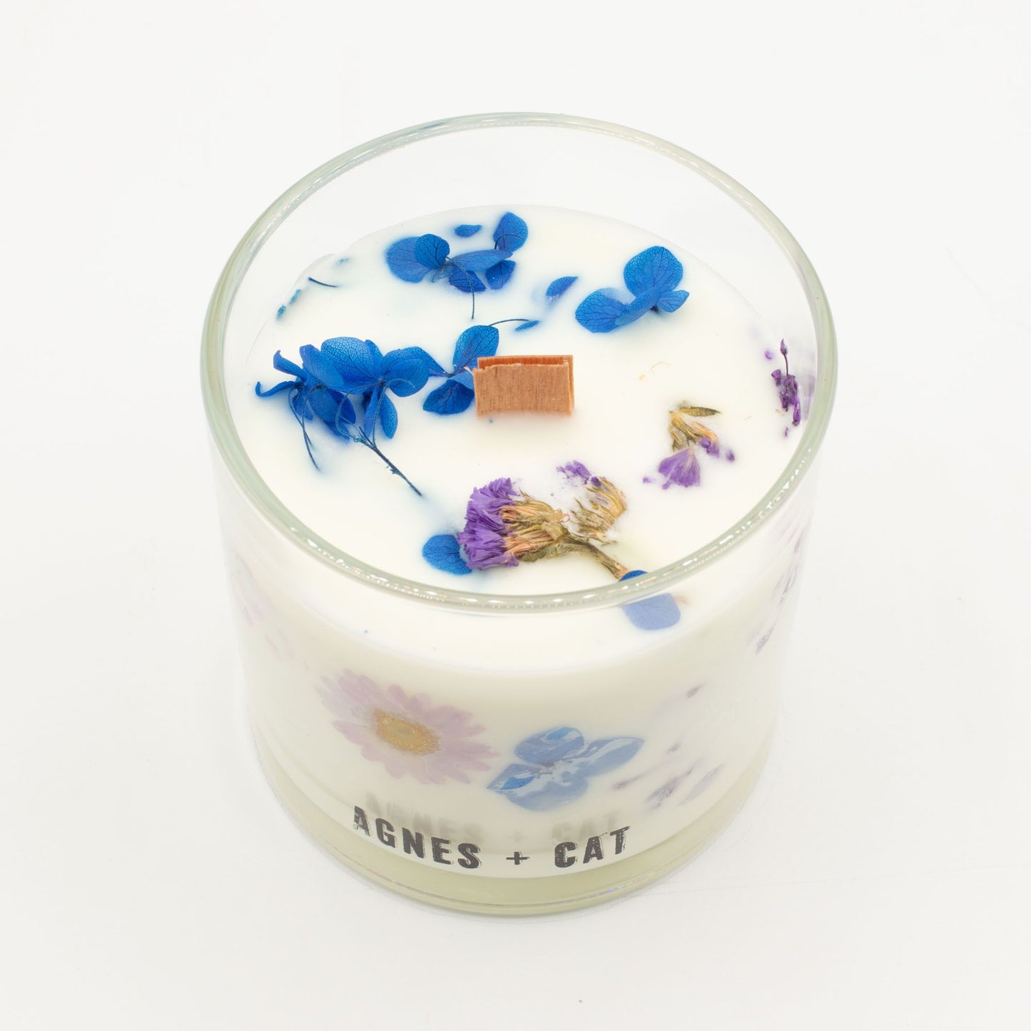 Vela aromática blanca con flores secas en azul y morado, en envase de vidrio con tapa de madera.