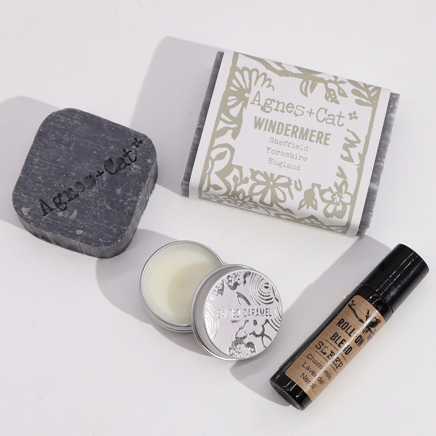 Set de regalo: jabón gris, bálsamo labial metálico y roll-on de aromaterapia sobre fondo blanco. Estilo artesanal.