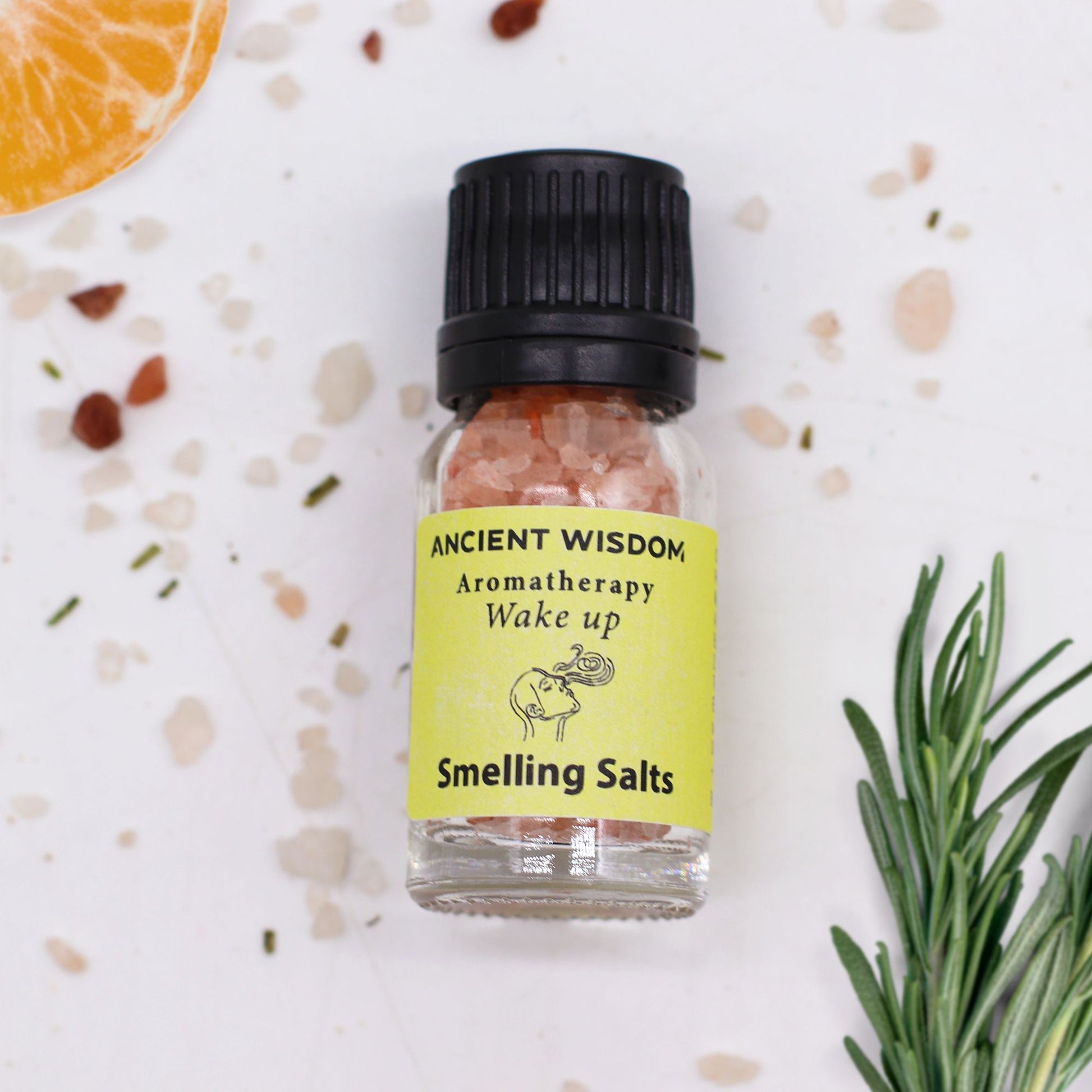 Frasco de sales aromáticas Ancient Wisdom, etiqueta amarilla, sales rosadas, rodeado de rodaja de naranja y romero.