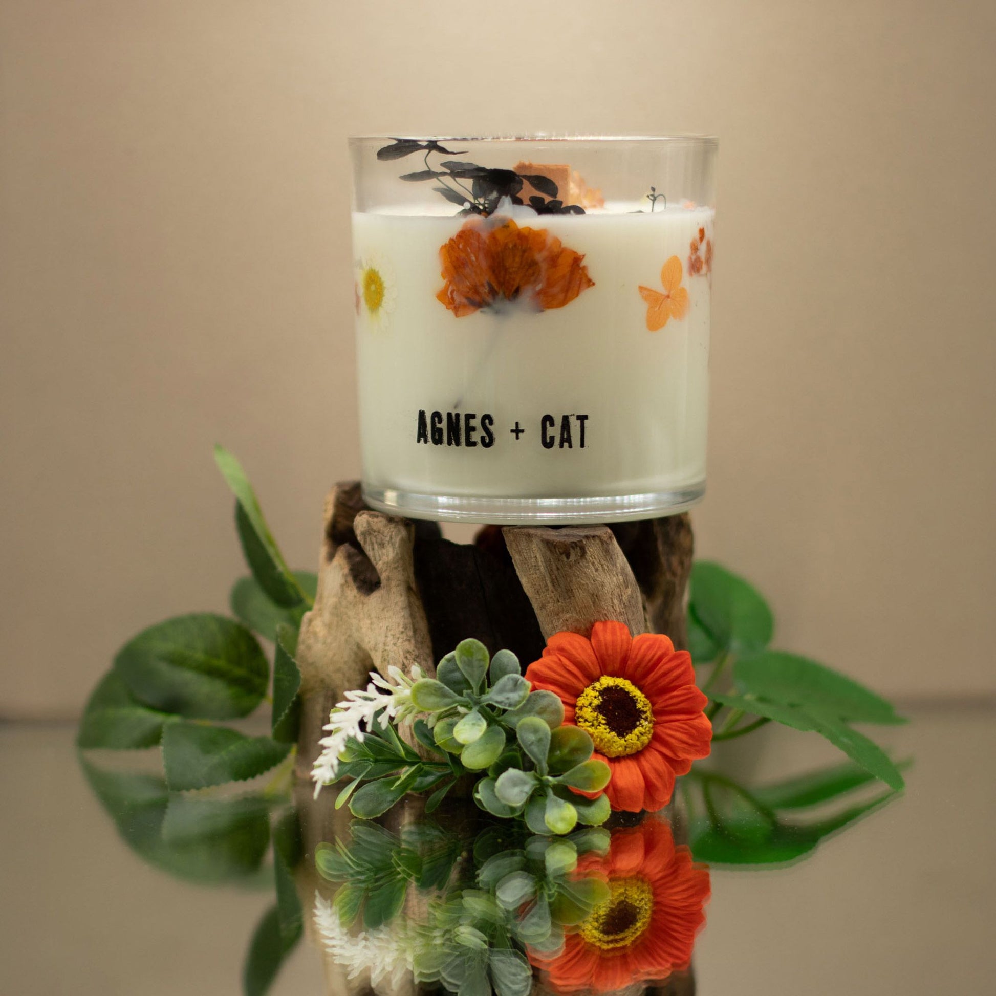 Vela aromática blanca con flores secas, en vaso de vidrio transparente, estilo bohemio, sobre soporte de madera.
