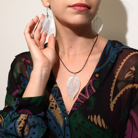 Collar y aretes plateados en forma de hoja, con cordón negro, luciendo elegante sobre blusa colorida.