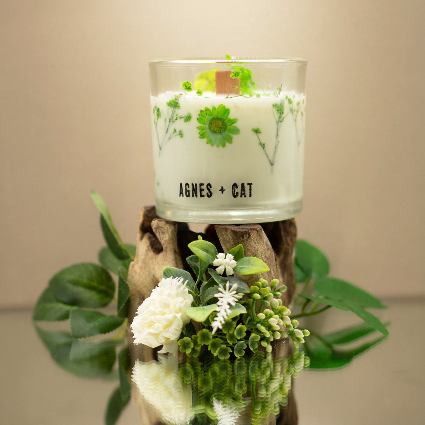 Vela aromática blanca con flores verdes, en vaso de cristal, sobre soporte de madera decorativo.