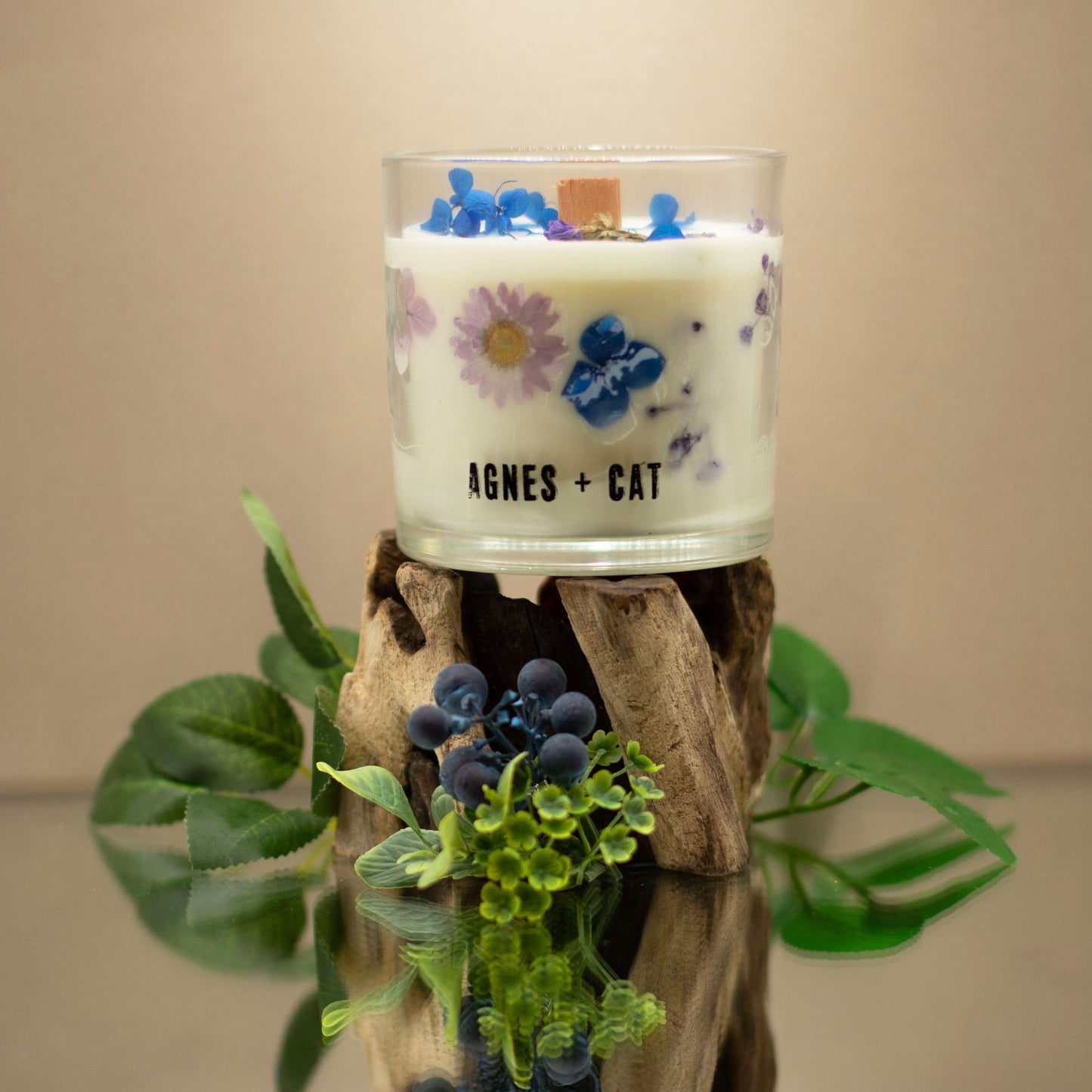 Vela aromática blanca con flores secas en vaso de vidrio, decorada sobre base de madera natural.