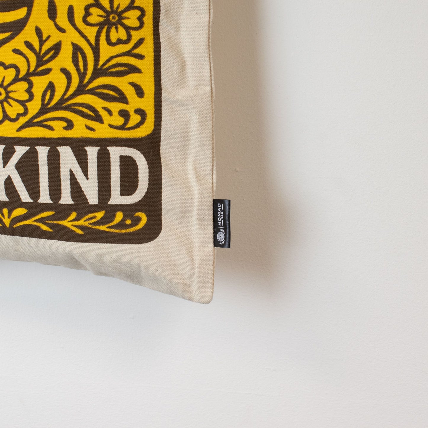 Cojín beige con diseño amarillento y marrón que dice "Be Kind", de tela suave y estampado artístico.