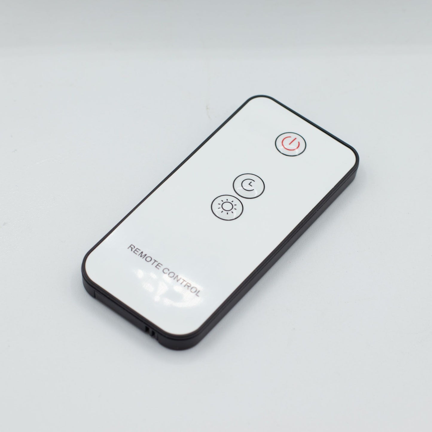 Mando a distancia blanco con borde negro, tres botones circulares y texto "REMOTE CONTROL" sobre fondo gris claro.