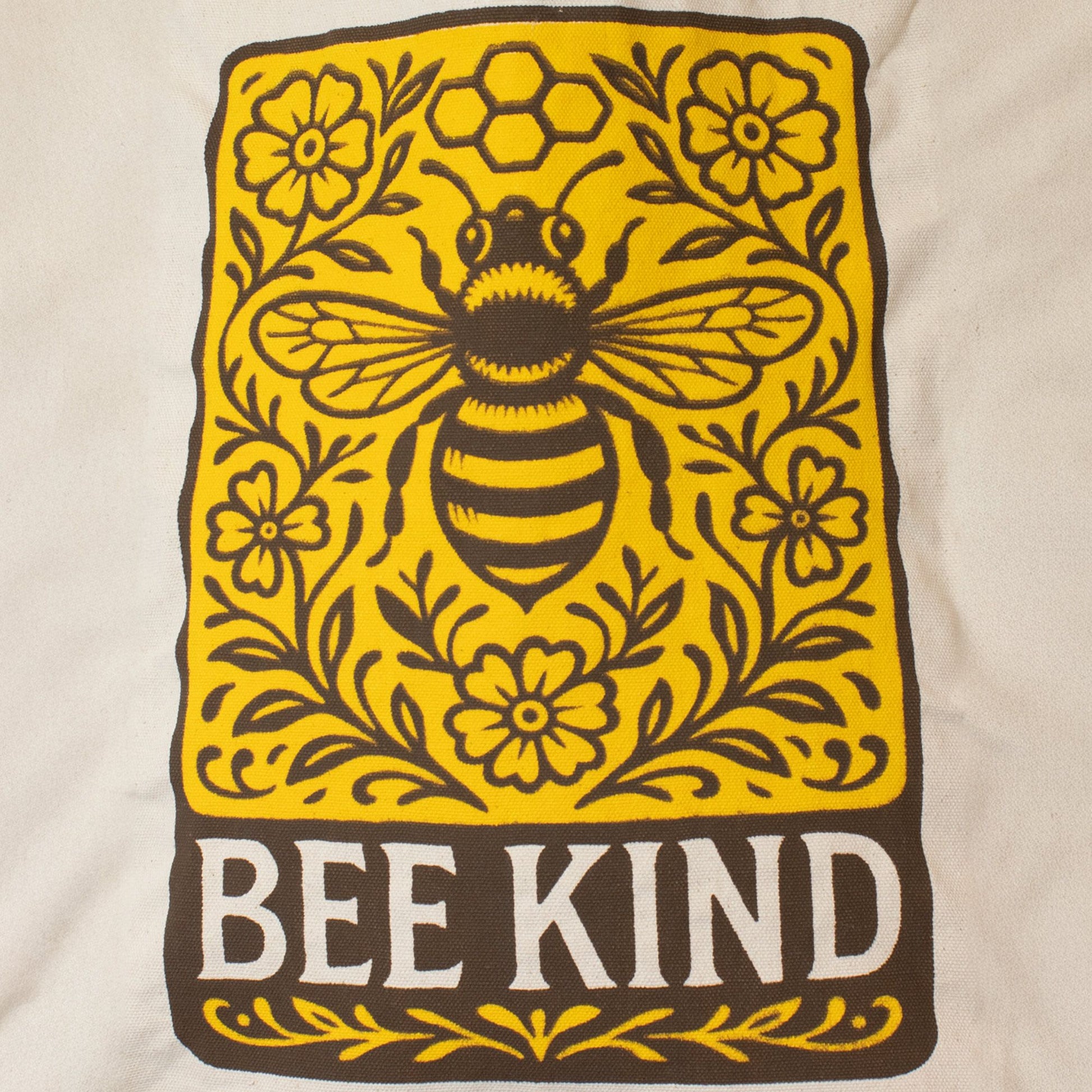 Diseño de bolsa de tela blanca con gráfico amarillo y marrón de abeja y flores, texto "BEE KIND".