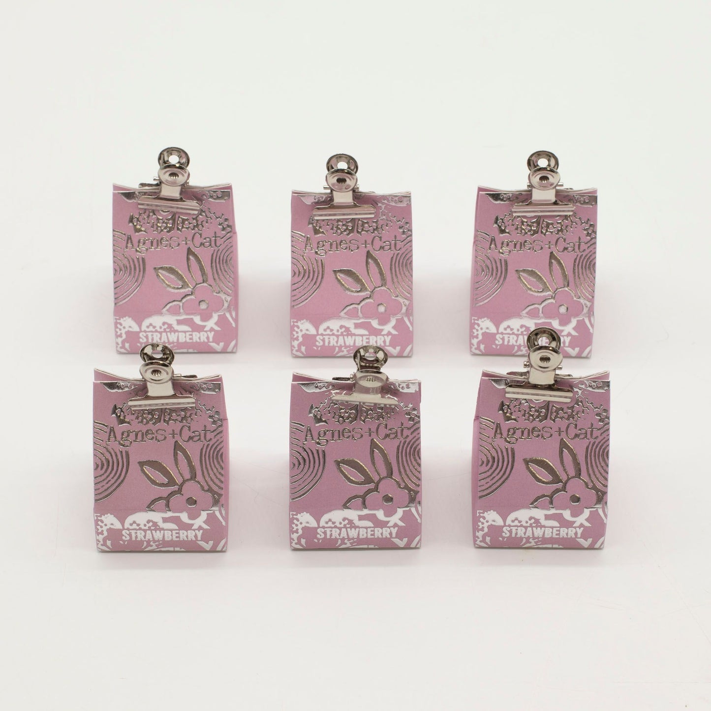 Seis bálsamos labiales Agnes + Cat de fresa en bolsas de papel rosa con pinzas metálicas sobre fondo blanco.