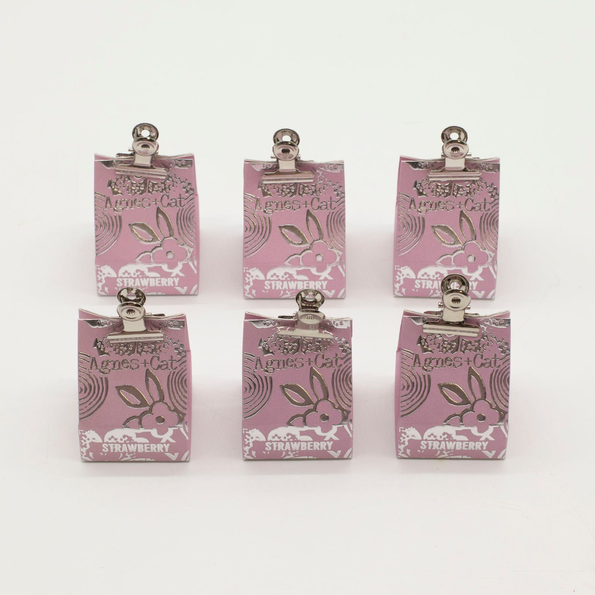 Seis bálsamos labiales Agnes + Cat de fresa en bolsas de papel rosa con pinzas metálicas sobre fondo blanco.