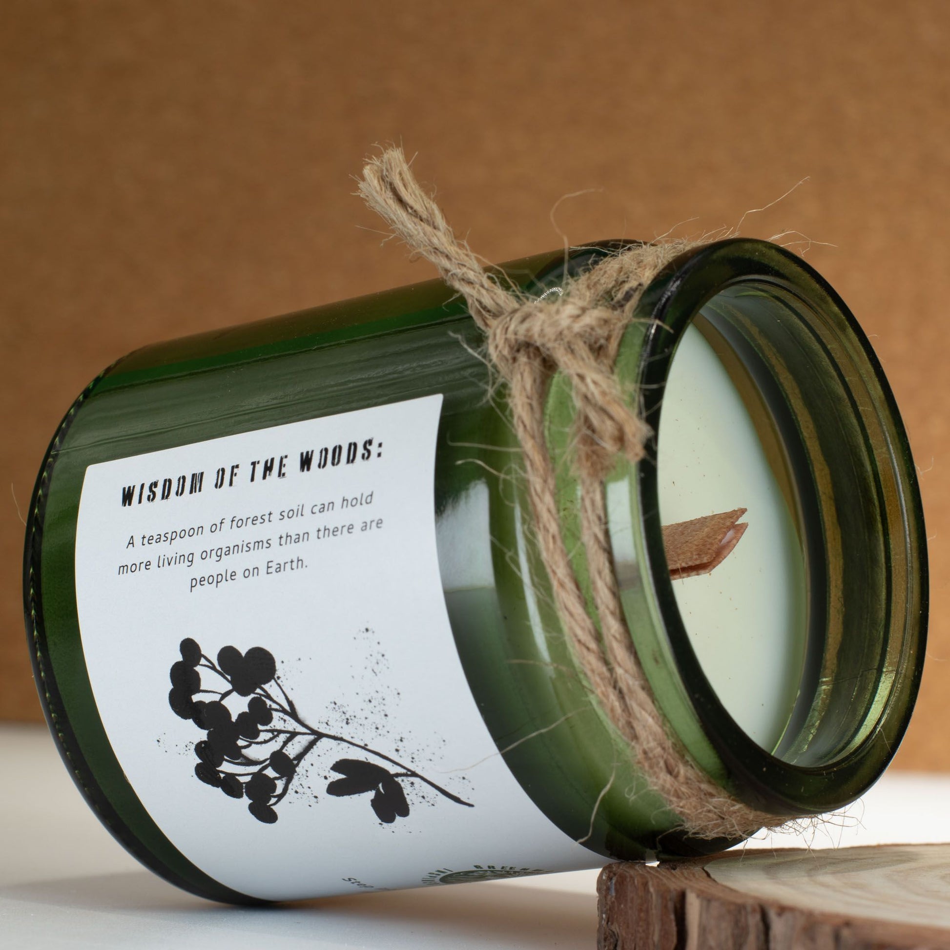 Vela verde de cristal con aroma a bosque, mecha de madera, etiqueta en blanco con texto sobre la vida en la tierra.