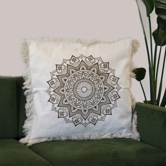Funda de cojín beige cuadrada con diseño de mandala en bronce y bordes decorados con flecos, sobre un sofá verde.