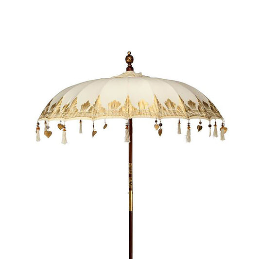 Sombrilla de patio estilo Bali en lona color crema con detalles dorados, flecos y poste de madera sobre fondo blanco