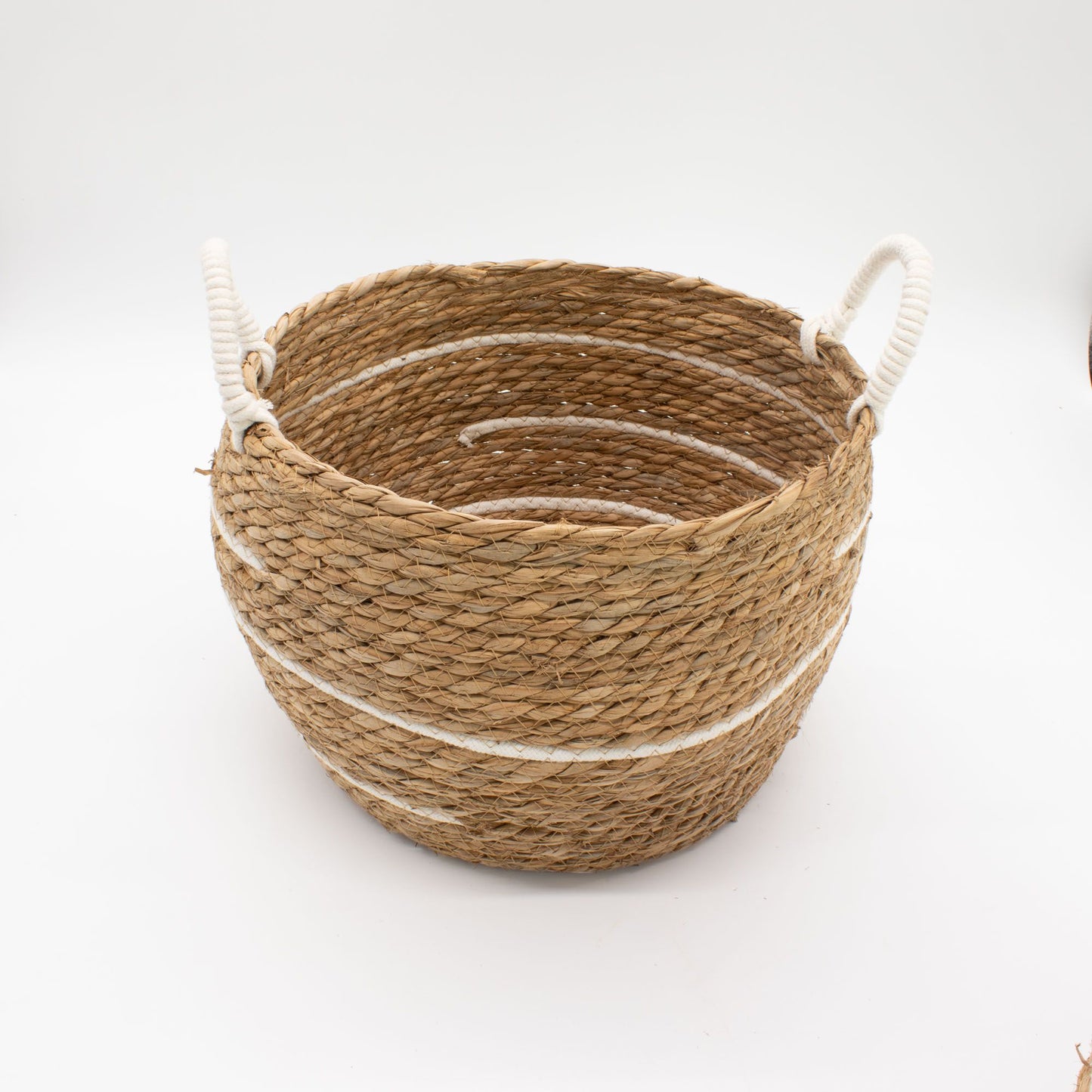 Cesta redonda de pasto marino trenzado con rayas de algodón blanco y asas laterales de cuerda, sobre fondo blanco.