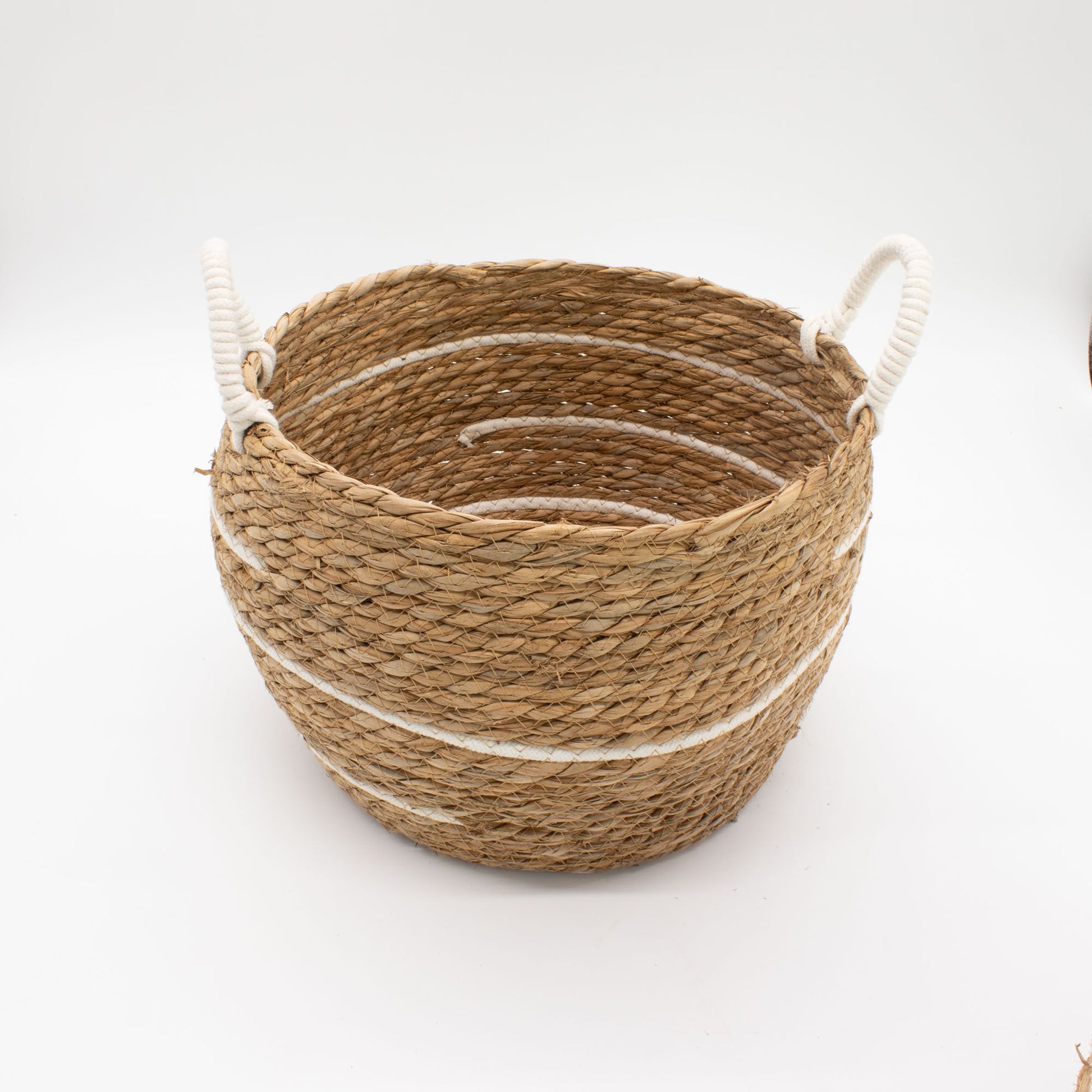 Cesta redonda de pasto marino trenzado con rayas de algodón blanco y asas laterales de cuerda, sobre fondo blanco.