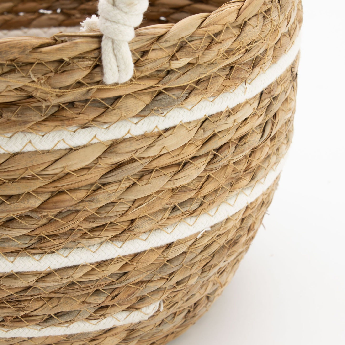 Cesta de pasto marino trenzado con rayas horizontales blancas de algodón y asa de cuerda sobre fondo blanco.