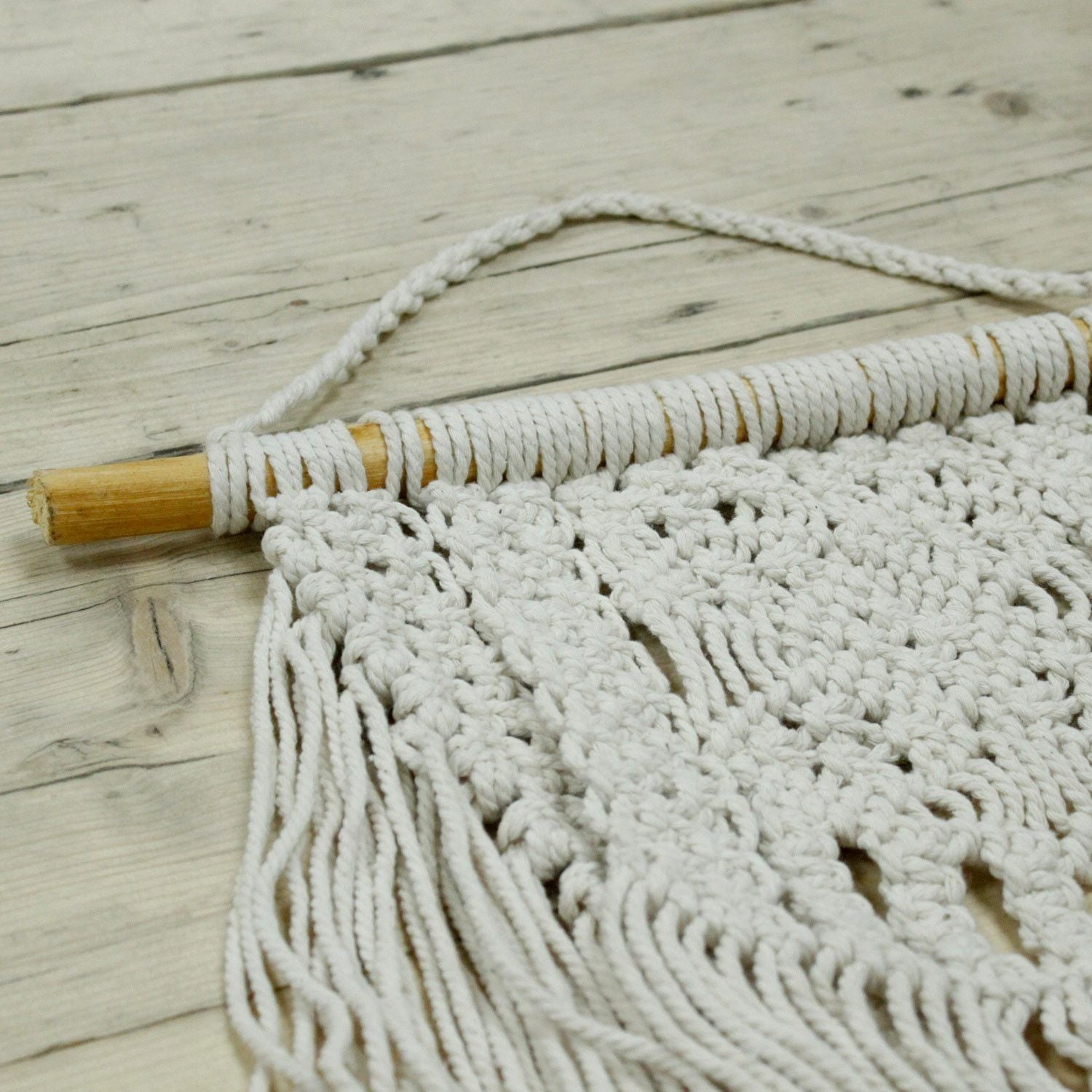 Mural de macramé blanco hecho a mano con barra de madera y flecos, ideal para decorar paredes.