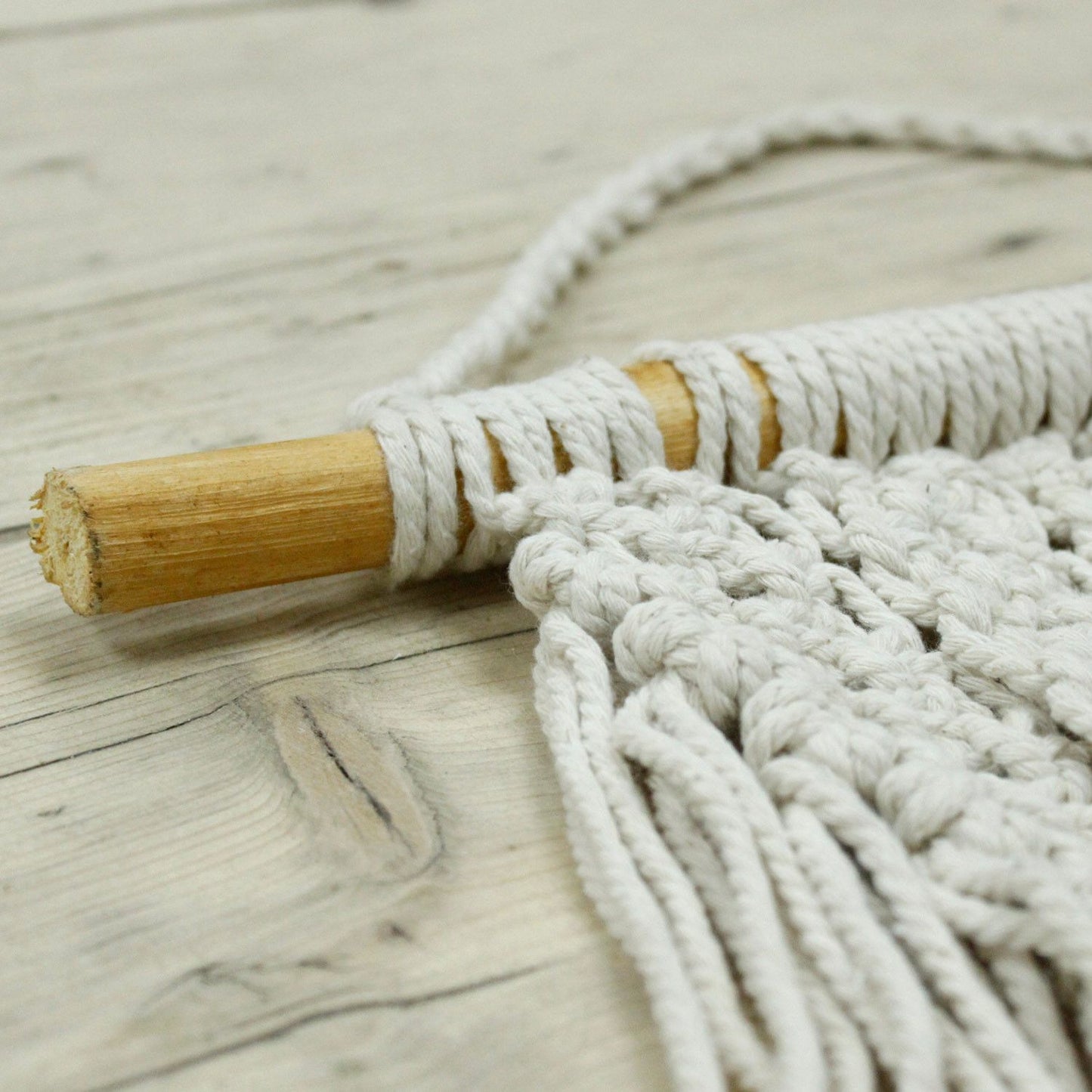 Cojín de macramé beige con barra de madera, textura suave y estilo bohemio para decoración del hogar.