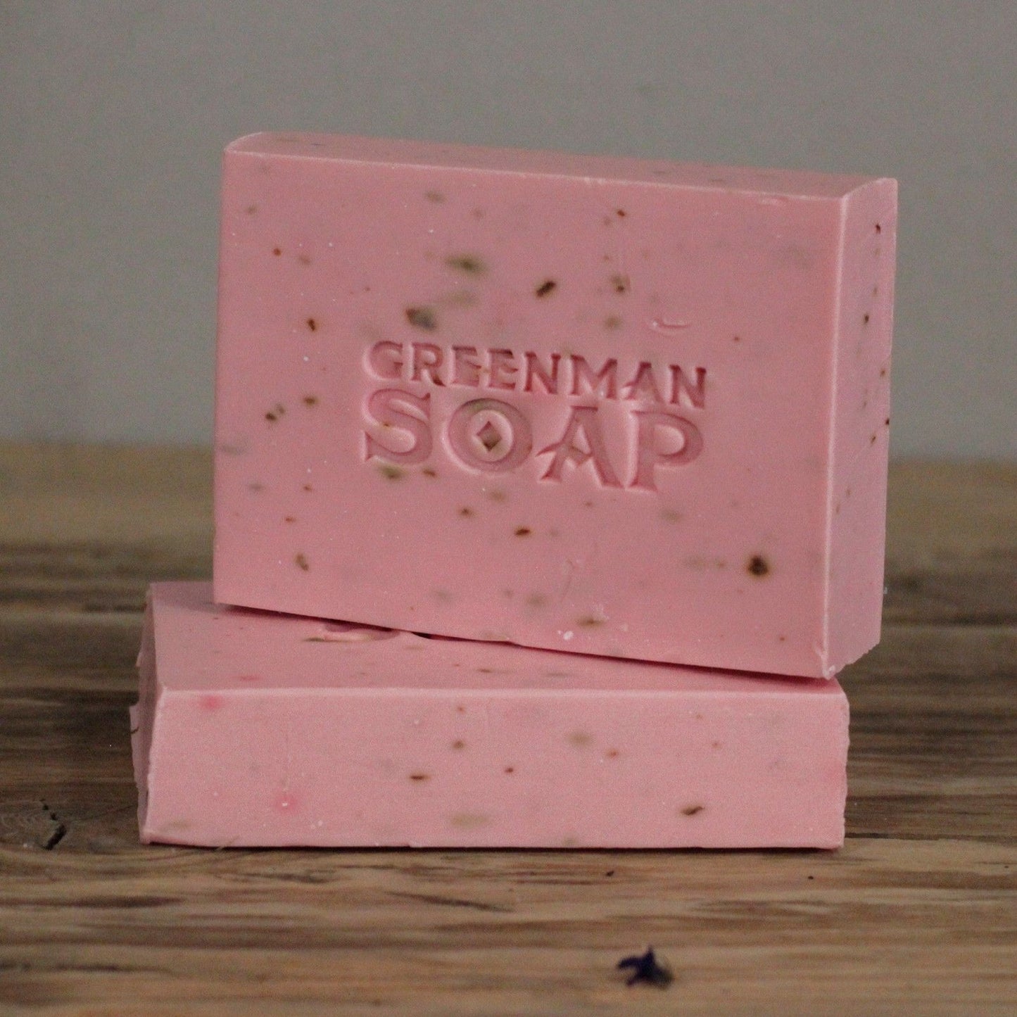 Dos pastillas de jabón rosa con motas, marcadas como "Greenman Soap", apiladas sobre una superficie de madera rústica.