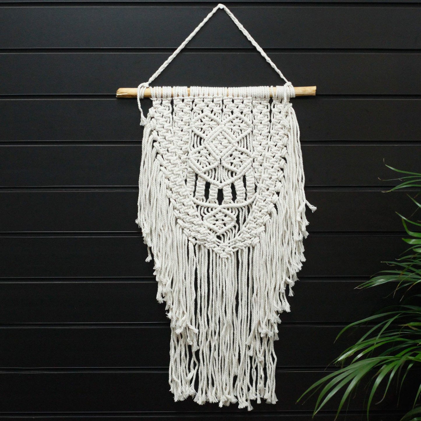 Macramé decorativo blanco con flecos en forma de diamante, colgado en una barra de madera. Ideal para interiores.