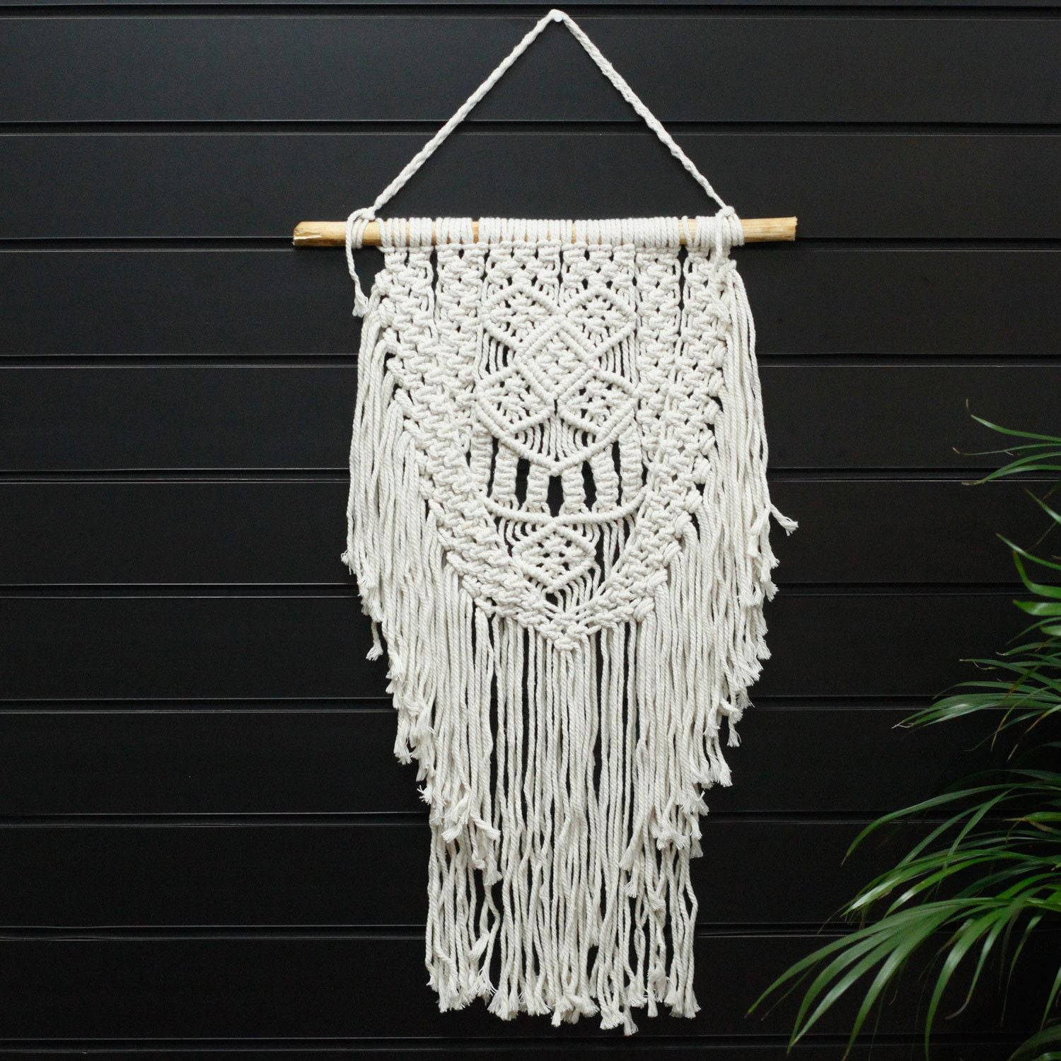 Macramé decorativo blanco con flecos en forma de diamante, colgado en una barra de madera. Ideal para interiores.