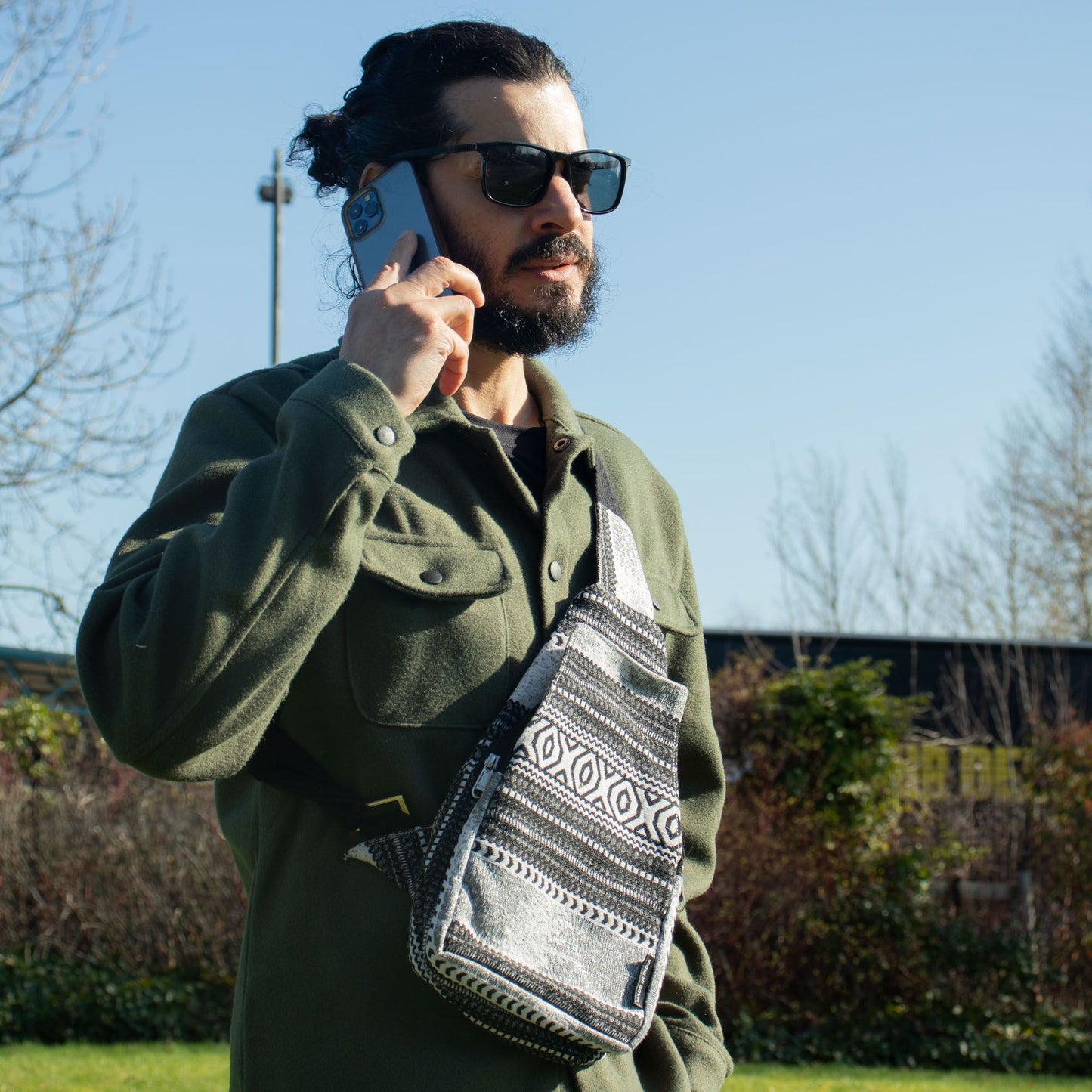 Hombre con bolsa cruzada gris con patrones, vestido con chaqueta verde y gafas de sol, hablando por teléfono.
