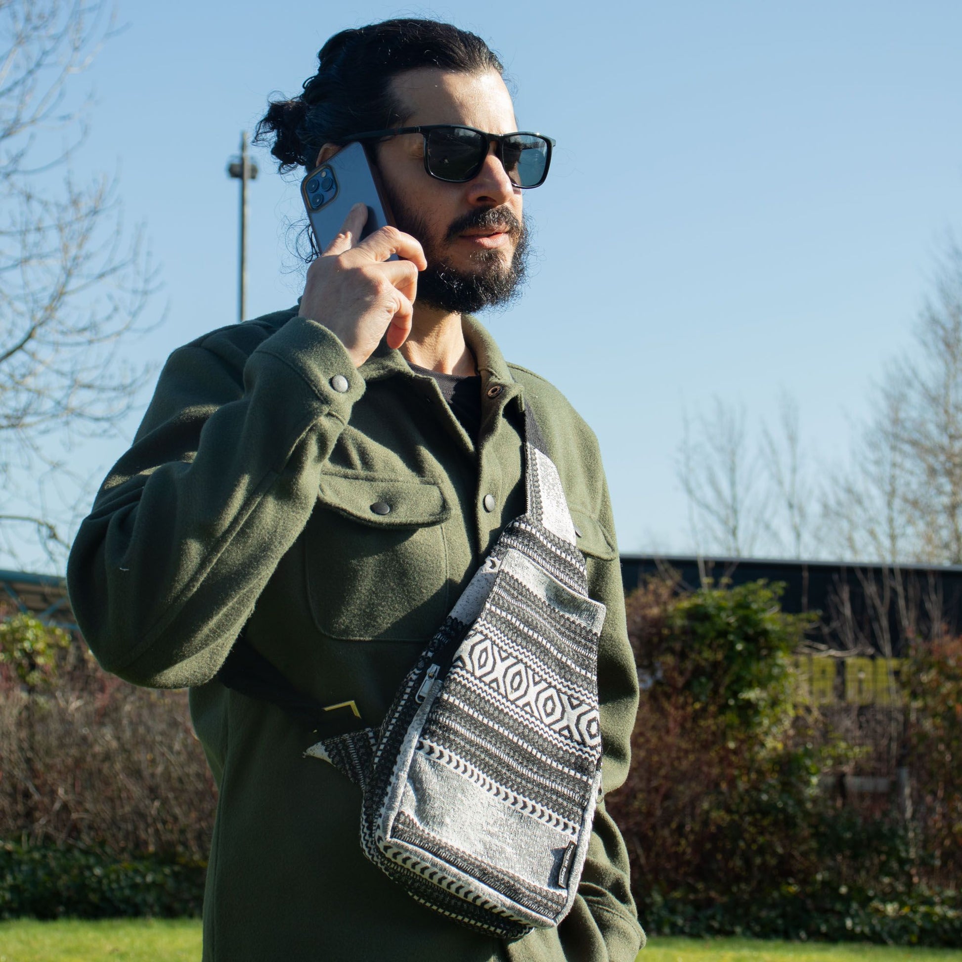 Hombre con bolsa cruzada gris con patrones, vestido con chaqueta verde y gafas de sol, hablando por teléfono.