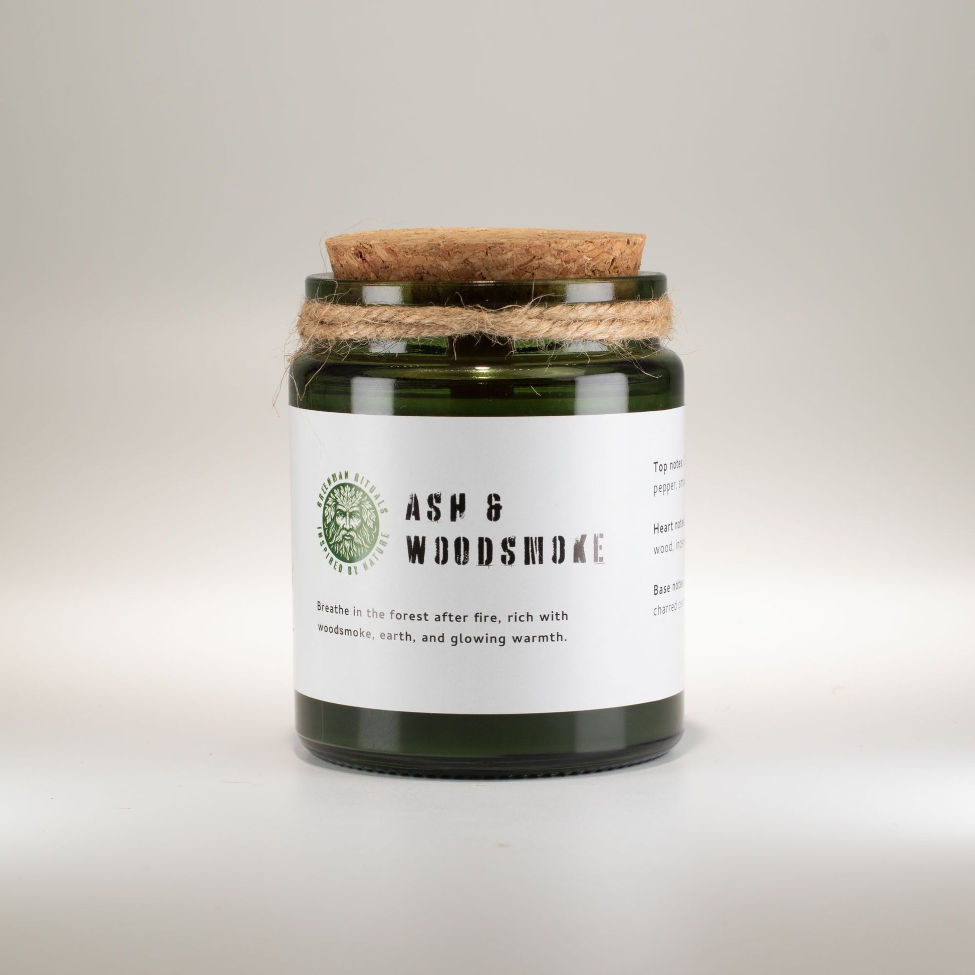 Vela de vidrio verde "Ash & Woodsmoke" con tapa de madera, aroma a tierra y humo de leña, estilo rústico.