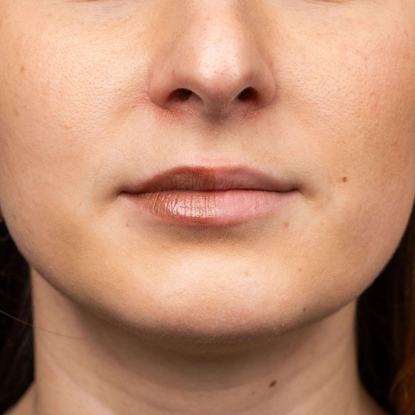 Close-up of lips with a glossy, hazelnut-nude tinted balm applied to the left side, contrasted with a bare right side.