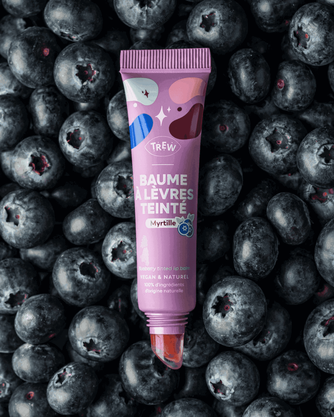 A lavender tube of blueberry tinted lip balm resting on a bed of fresh, dark blue blueberries.