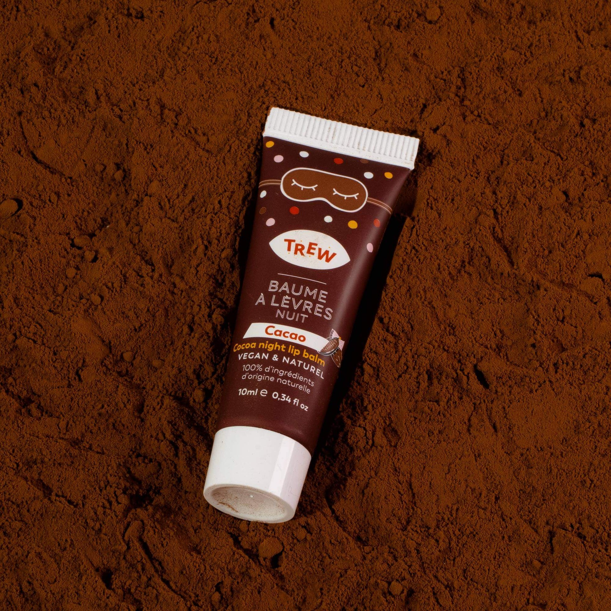A brown cocoa night lip balm tube with a sleep mask icon, resting on a bed of dark cocoa powder.